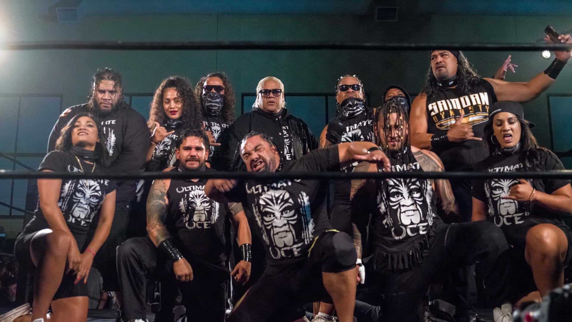 Major League Wrestler 雅各布·法圖 安奧亞家族 壁紙 — 一個兇猛的摔角派系自信地在擂台上擺姿勢，穿著搭配的黑色襯衫，上面有部落面具標誌。