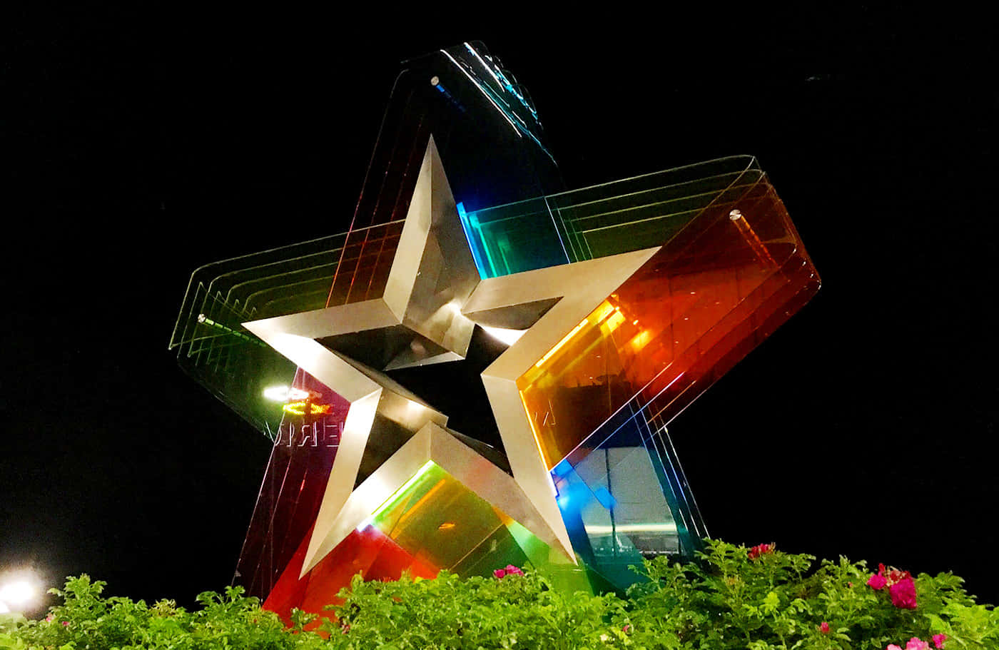 Mall of America購物天堂 壁紙 — 夜間發光的彩虹星星雕塑，在黑暗的天空和下方的綠色樹葉襯托下呈現鮮豔色彩。