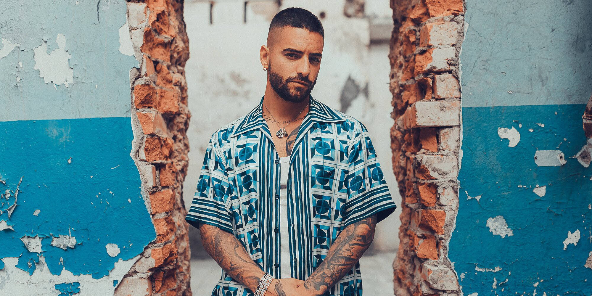 Maluma《#7DJ (7 Días en Jamaica)》專輯拍攝 壁紙 — 一個有紋身和鬍鬚的男人站在破爛的磚牆之間，背景是風化的藍白牆壁。