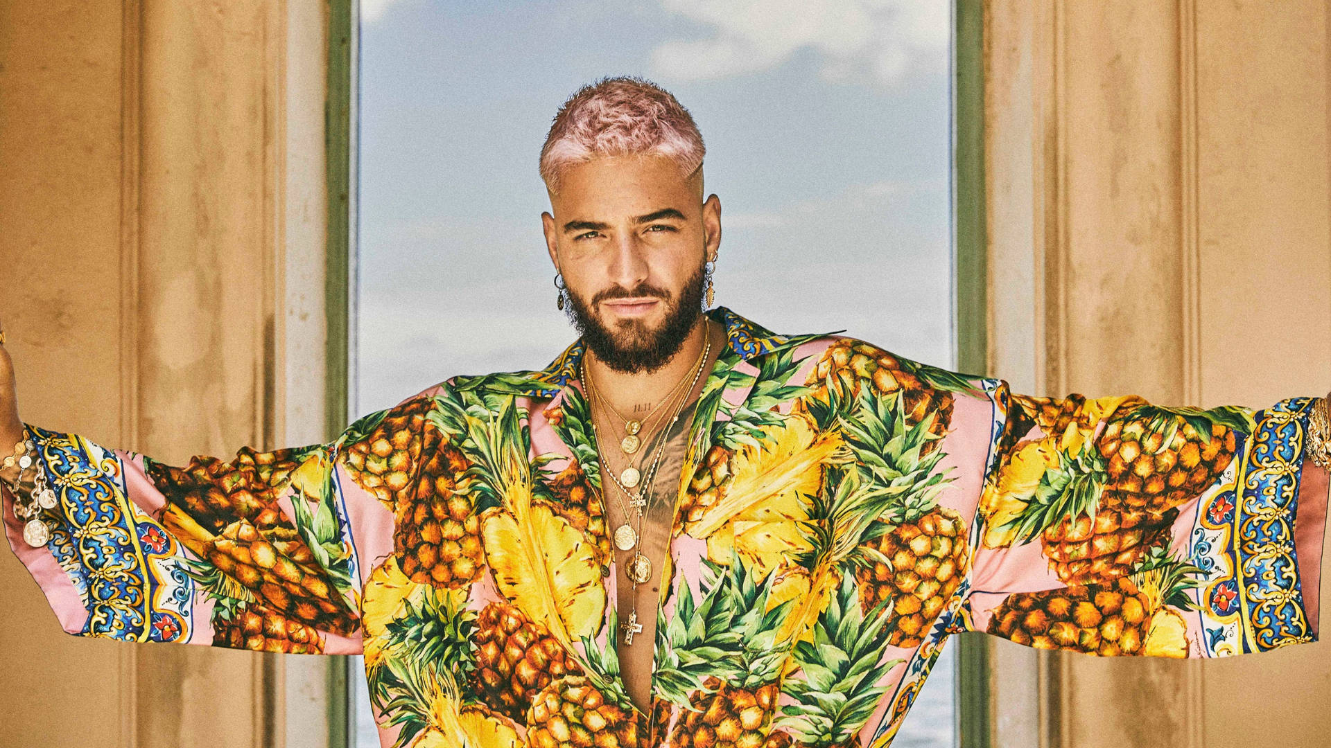 Maluma 英國GQ時尚拍攝 壁紙 — 一個粉紅色頭髮和鬍鬚的男人穿著鮮豔的菠蘿印花長袍，站在窗前，雙臂伸展。