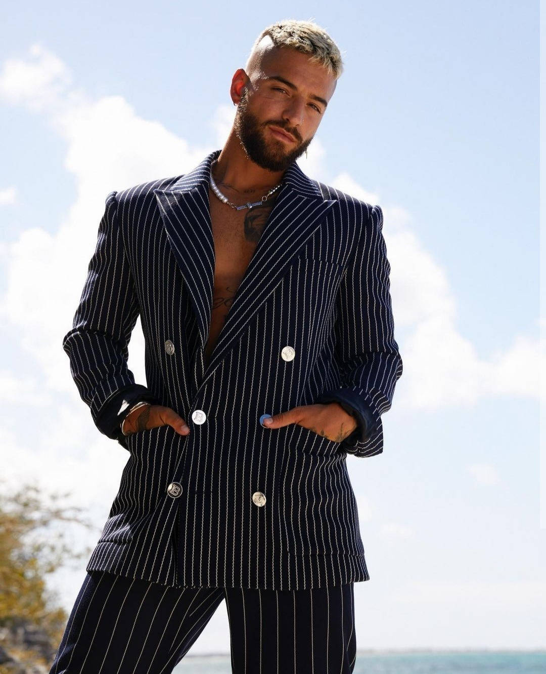 Maluma 西班牙《Cosmopolitan》封面拍攝 壁紙 — 一位時尚男士身穿海軍藍細條紋西裝，雙手插袋，自信地站在戶外，背景是明亮的藍天。