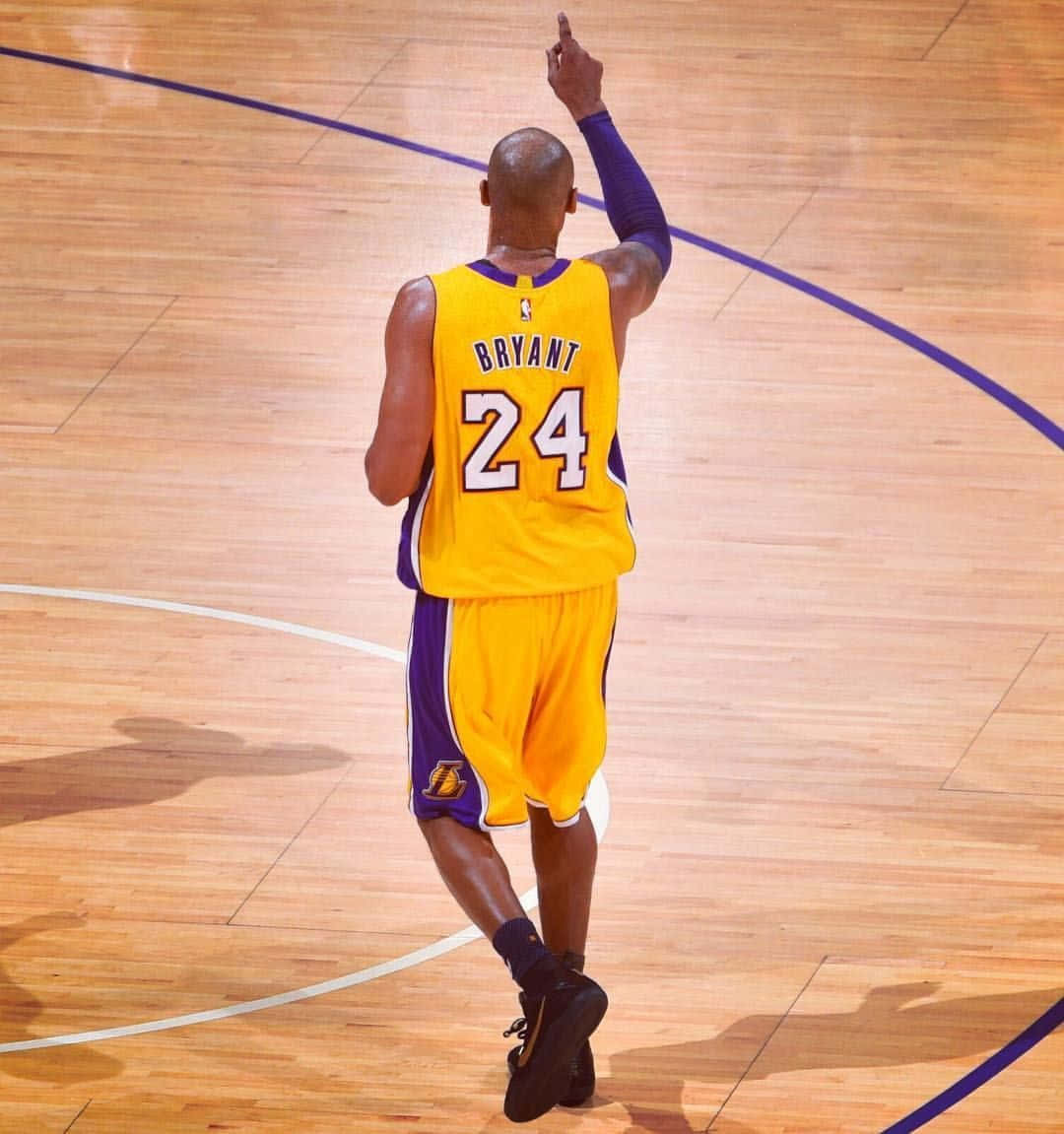 科比·布萊恩特的傳承永存 — #MambaOut 壁紙 — 一名身穿黃色和紫色球衣、背號24的籃球運動員站在木地板上向上指。