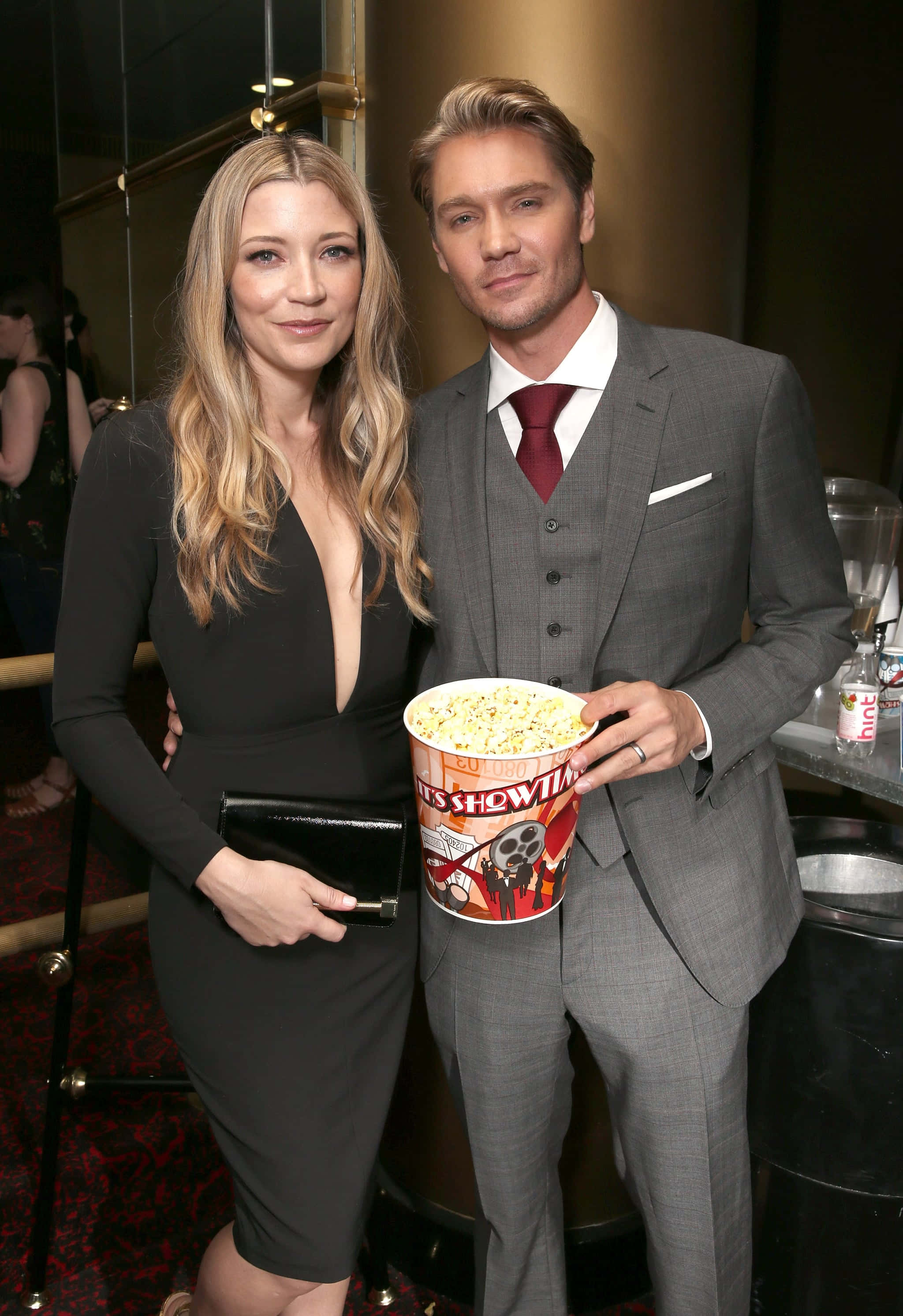 Sarah Roemer 桌布 壁紙 — 一位身穿灰色西裝的男士和一位身穿黑色連衣裙的女士在一個華麗的活動中一起擺姿勢，拿著一個大爆米花桶。