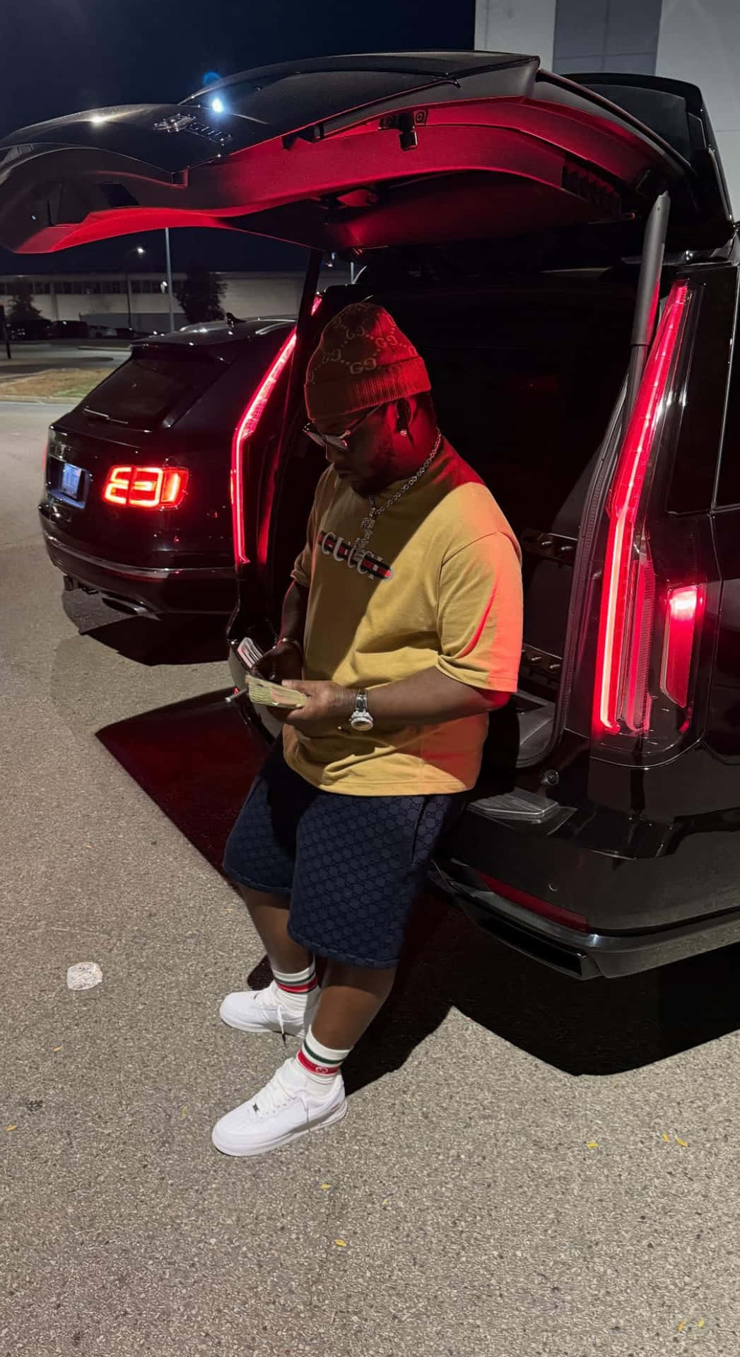  壁紙 — 一名身穿黃色襯衫和冷帽的男子，在夜晚靠著一輛黑色SUV打開的尾箱數錢。