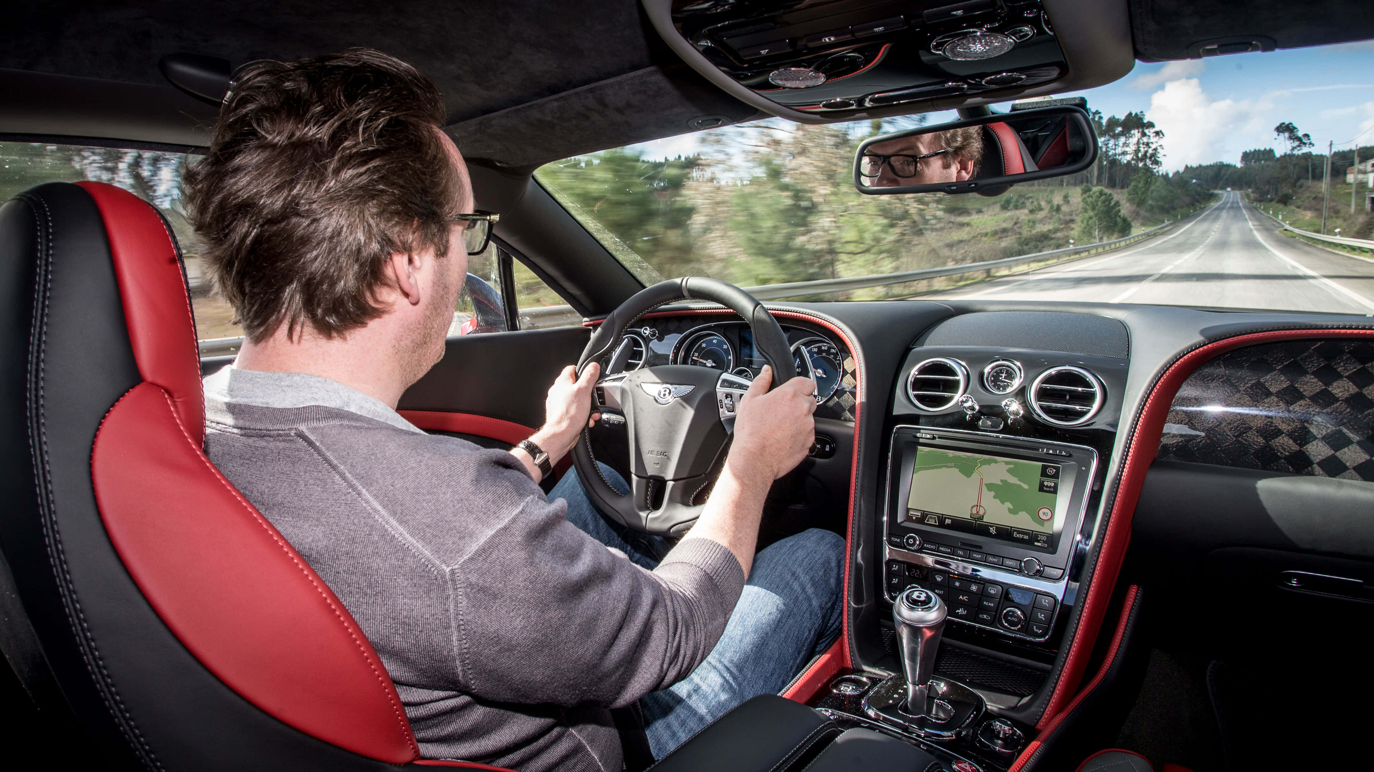 Bentley Continental Supersports 桌布 壁紙 — 一名男子駕駛一輛配有紅色皮革座椅的豪華汽車，沿著蜿蜒的道路穿過綠色的山丘，天空晴朗。