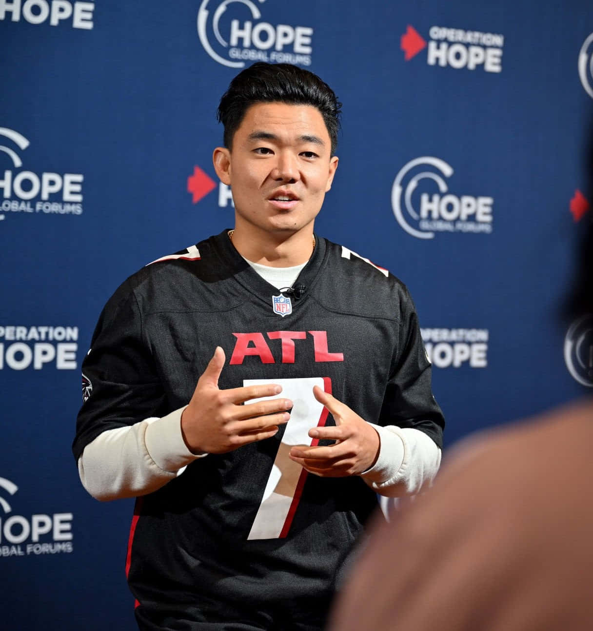  壁紙 — 一名身穿亞特蘭大獵鷹球衣的男子在Hope Global Forums活動中，在品牌藍色背景前說話並做手勢。