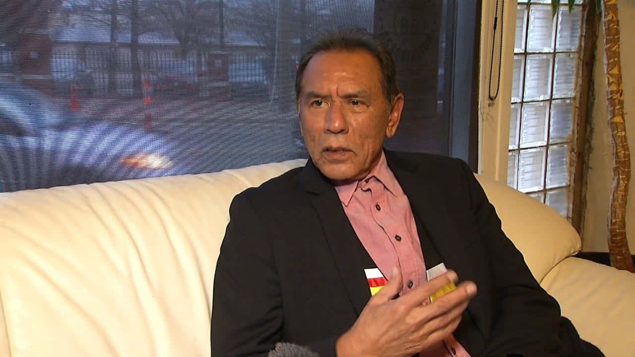  壁紙 — 一名身穿黑色西裝和粉紅色襯衫的男子坐在白色沙發上，手持黃色物體，直視鏡頭。