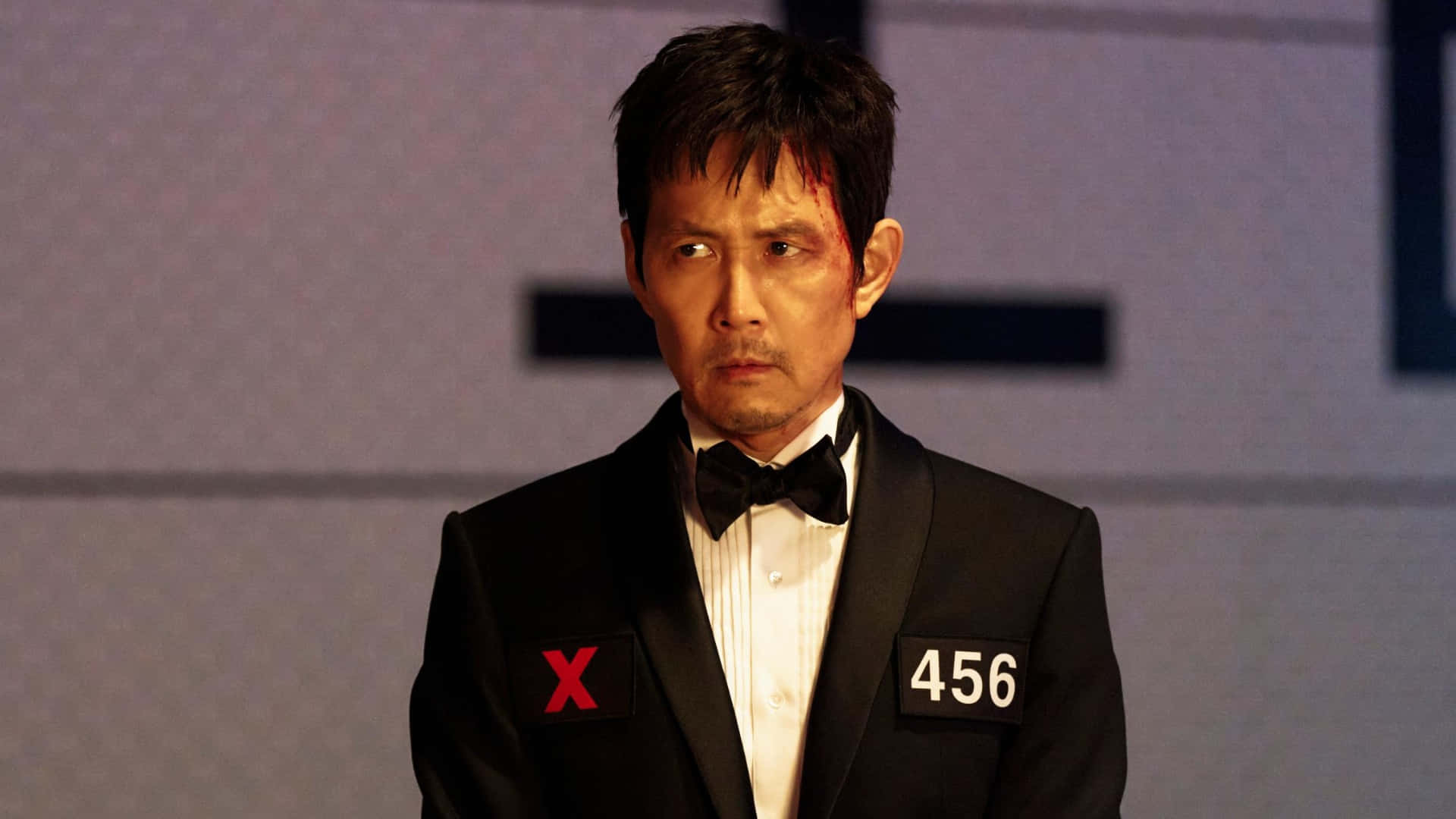 壁紙 — 一名身穿黑色燕尾服的男子，外套上有紅色X和數字456，帶著瘀傷的臉，在純色背景前專注地凝視。