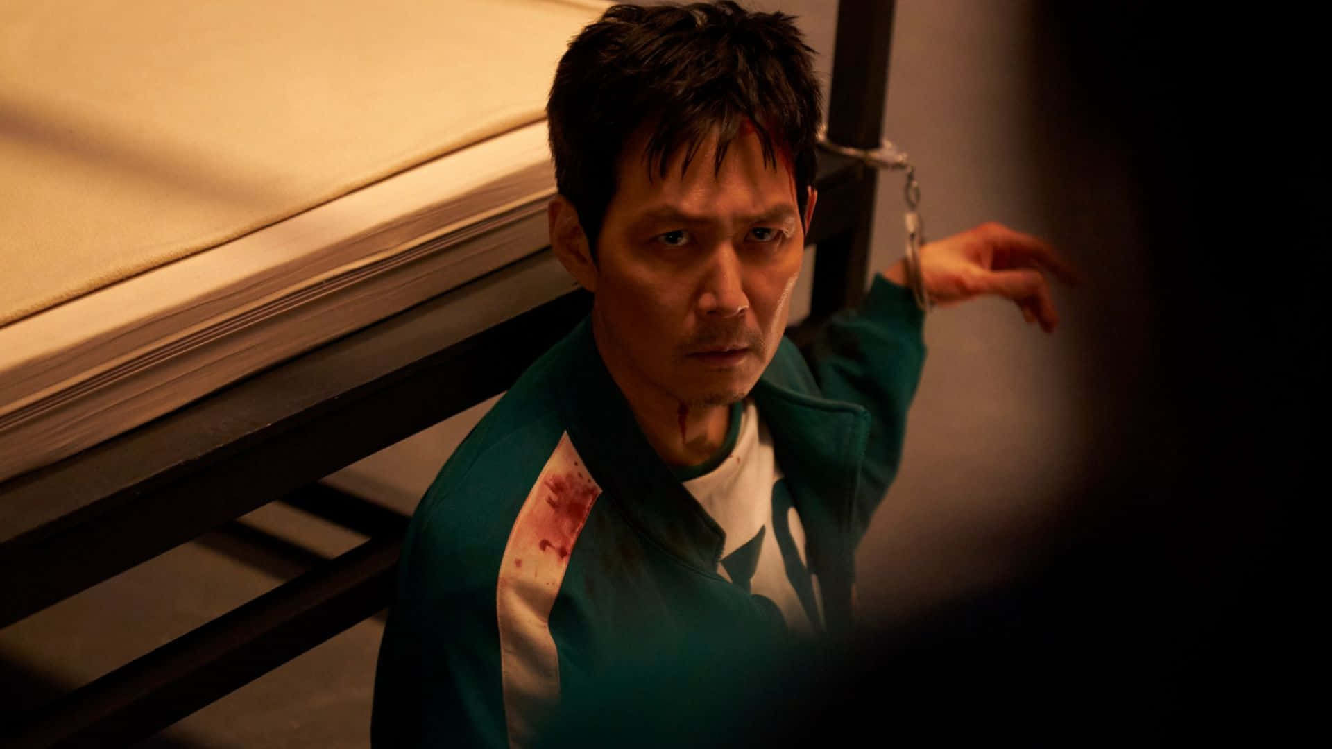  壁紙 — 一個身穿沾血綠色運動服的男人在昏暗的房間裡被銬在雙層床上，眼神凌厲。