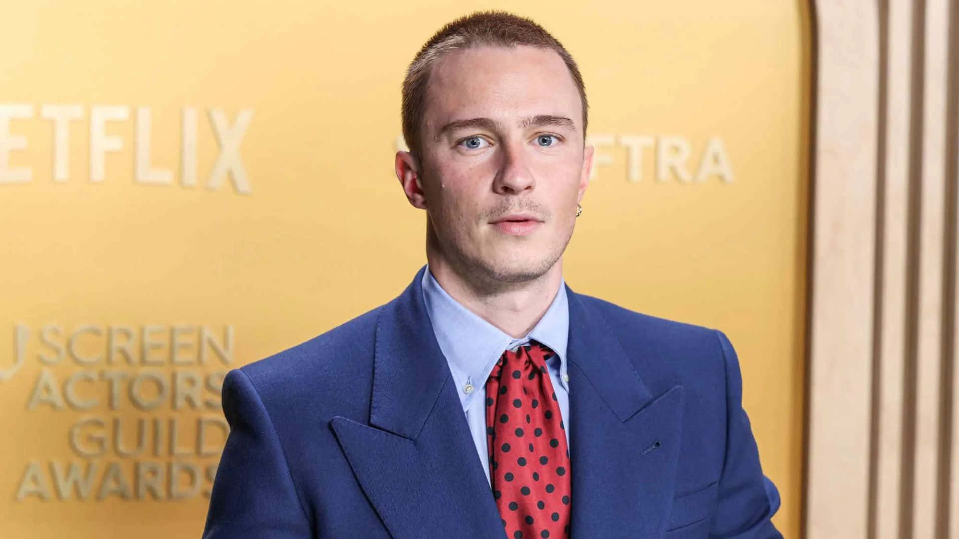  壁紙 — 一位身穿藍色西裝、紅色圓點領帶的男子自信地站在金色Netflix SAG Awards背景前。