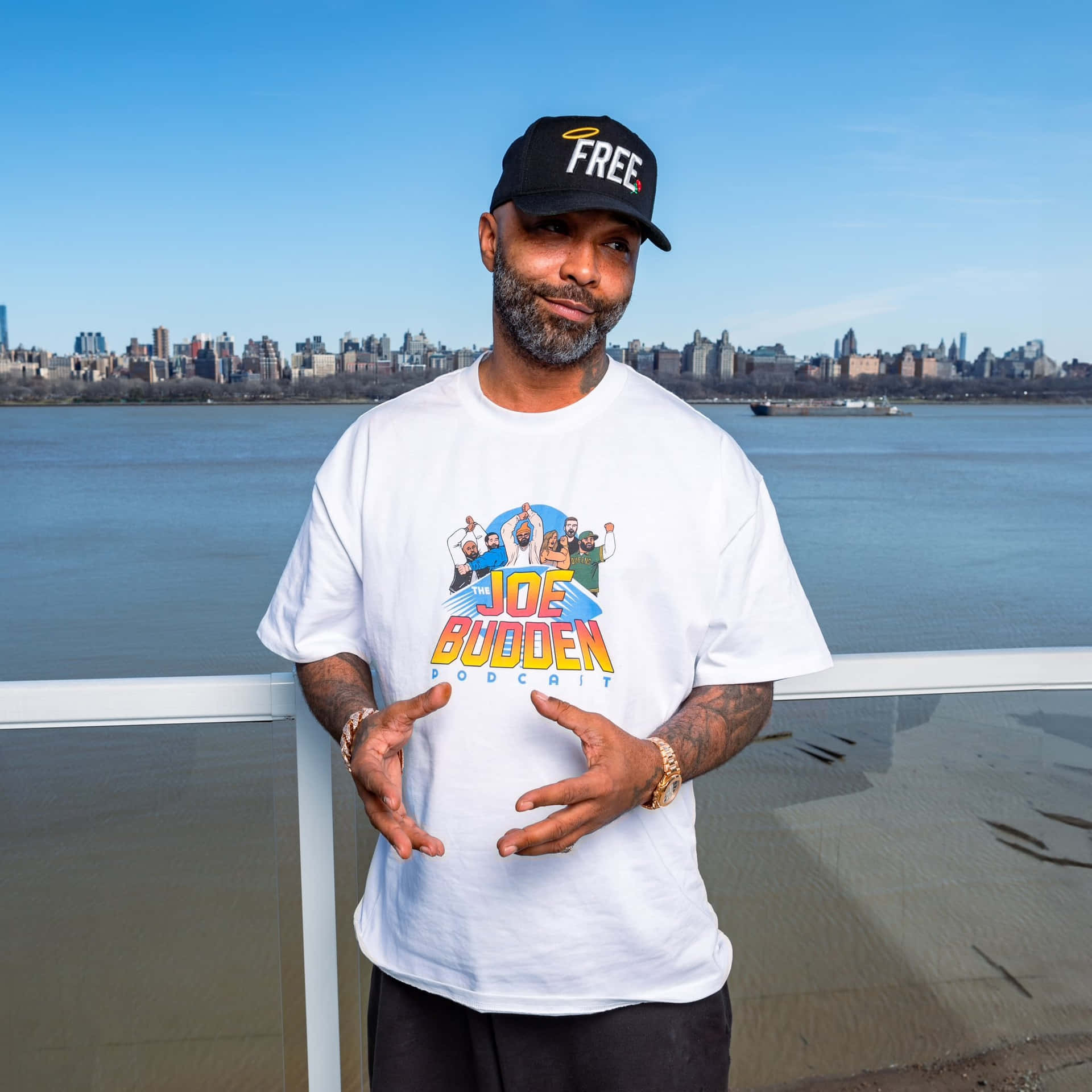  壁紙 — 一名戴著「FREE」帽子和穿著Joe Budden播客T恤的男子站在海濱陽台上，身後是城市天際線。