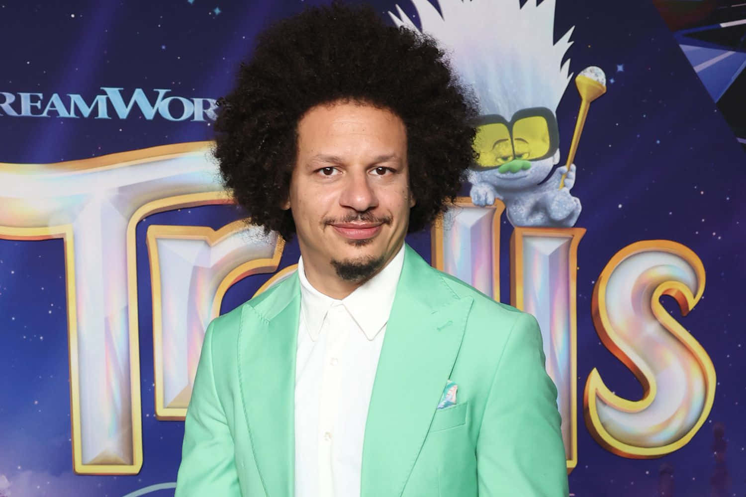  壁紙 — 一名捲髮男子身穿薄荷綠色西裝，站在帶有卡通角色的DreamWorks Trolls宣傳背景前。