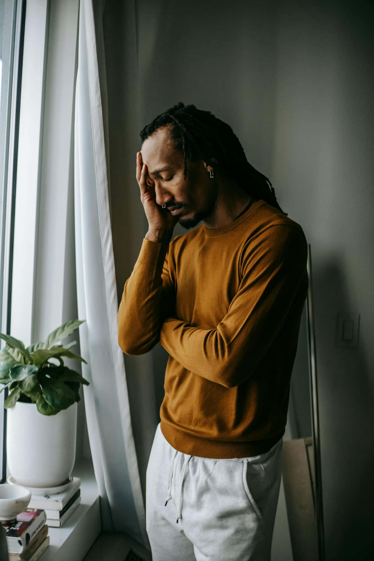  壁紙 — 一名身穿橙色毛衣的男子站在窗邊，雙手抱頭，看似痛苦或陷入沉思。