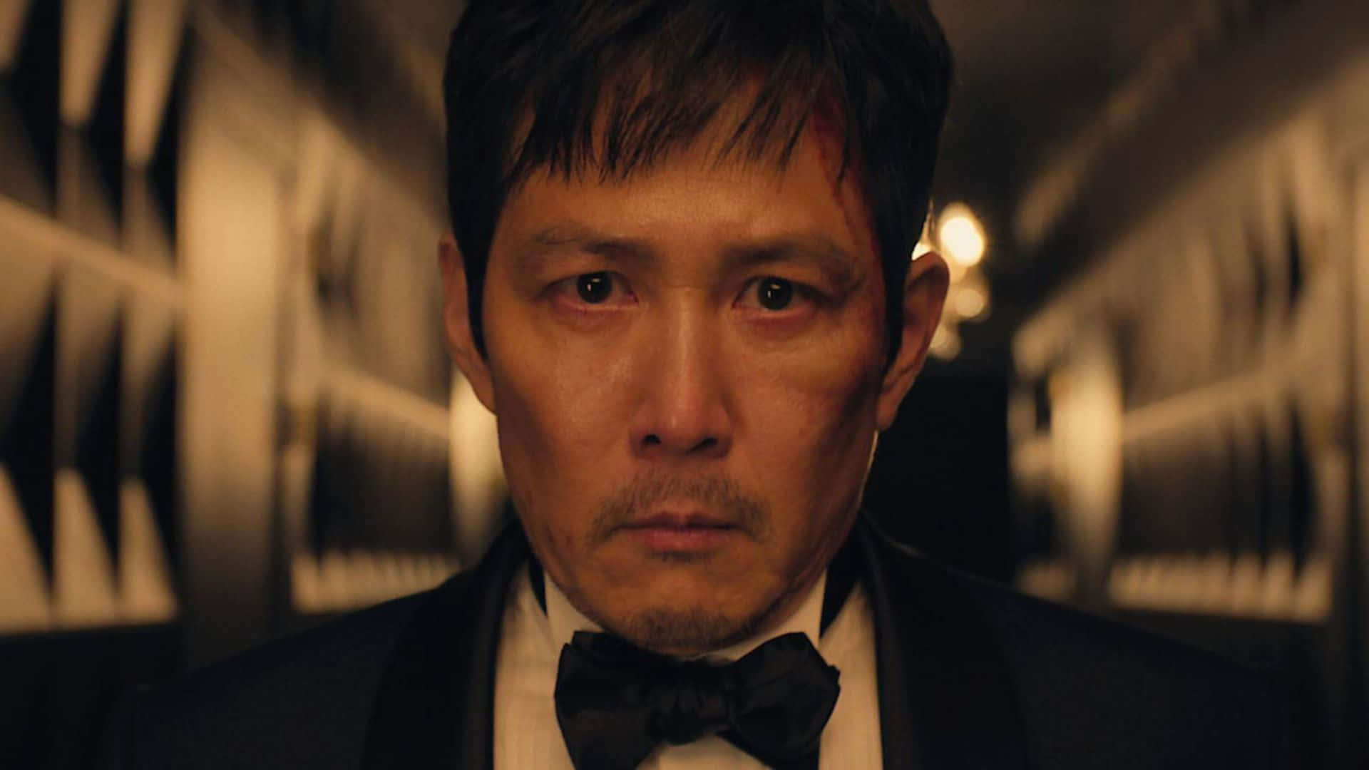  壁紙 — 一名身穿燕尾服的男人，沿著一條光線昏暗、帶有幾何圖案牆紙和溫暖發光燈光的走廊，凝視著前方。