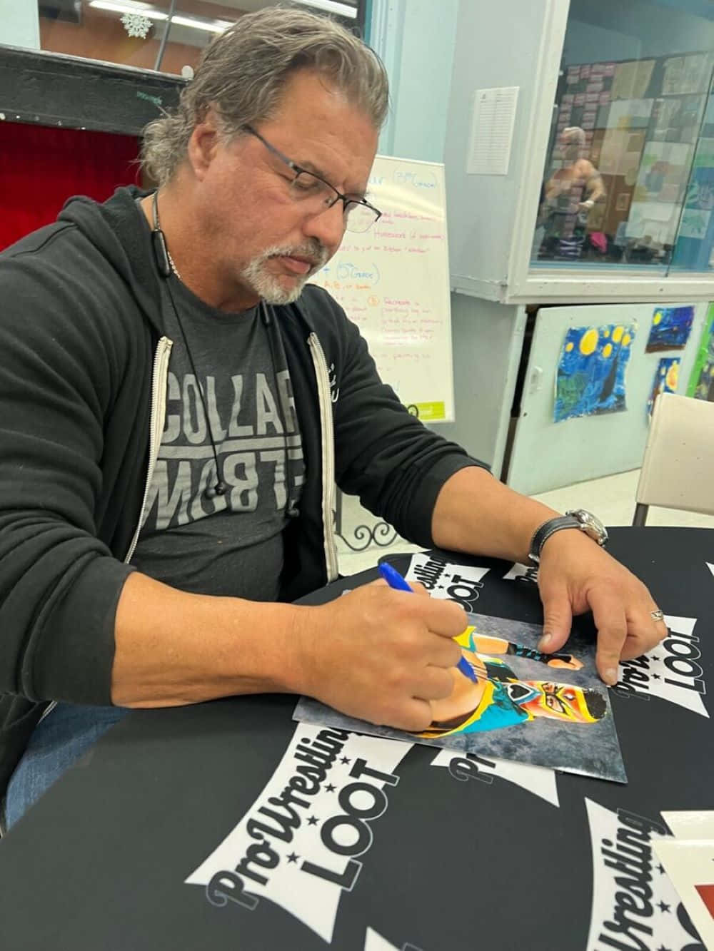  壁紙 — 一名戴眼鏡、灰白頭髮的男子在室內的ProWrestlingLOOT桌上簽署一張色彩繽紛的摔角海報。