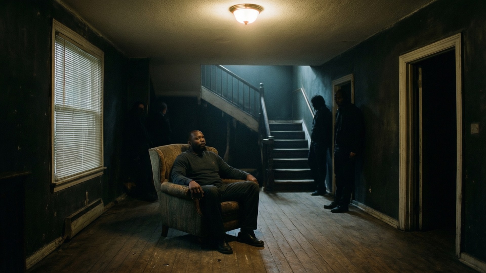 get out 桌布 壁紙 — 一名男子獨自坐在昏暗的走廊中，陰影人物在單一吊燈下的樓梯附近若隱若現。