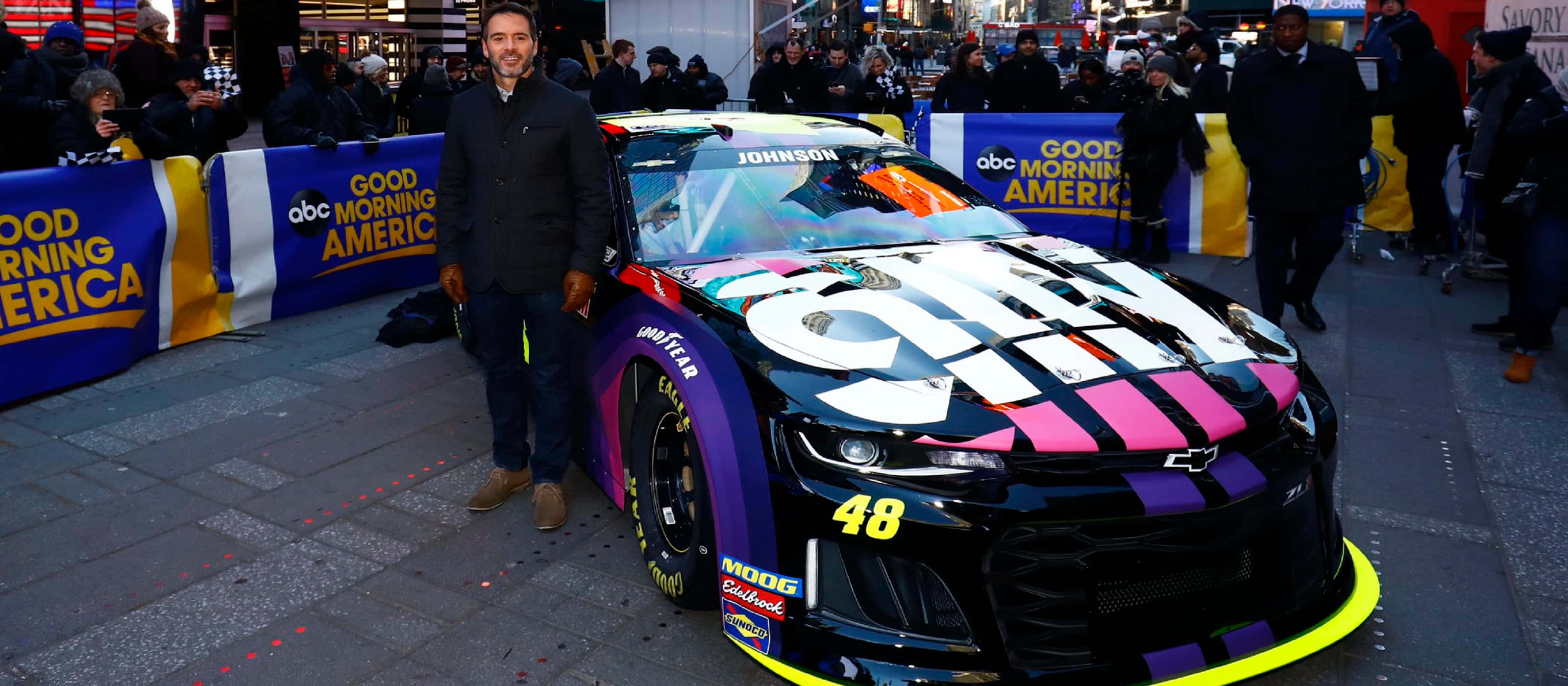 Jimmie Johnson 桌布 壁紙 — 一名男子站在一輛充滿活力的黑色賽車旁，賽車帶有粉紅、紫色和黃色點綴，在繁忙的城市活動中。