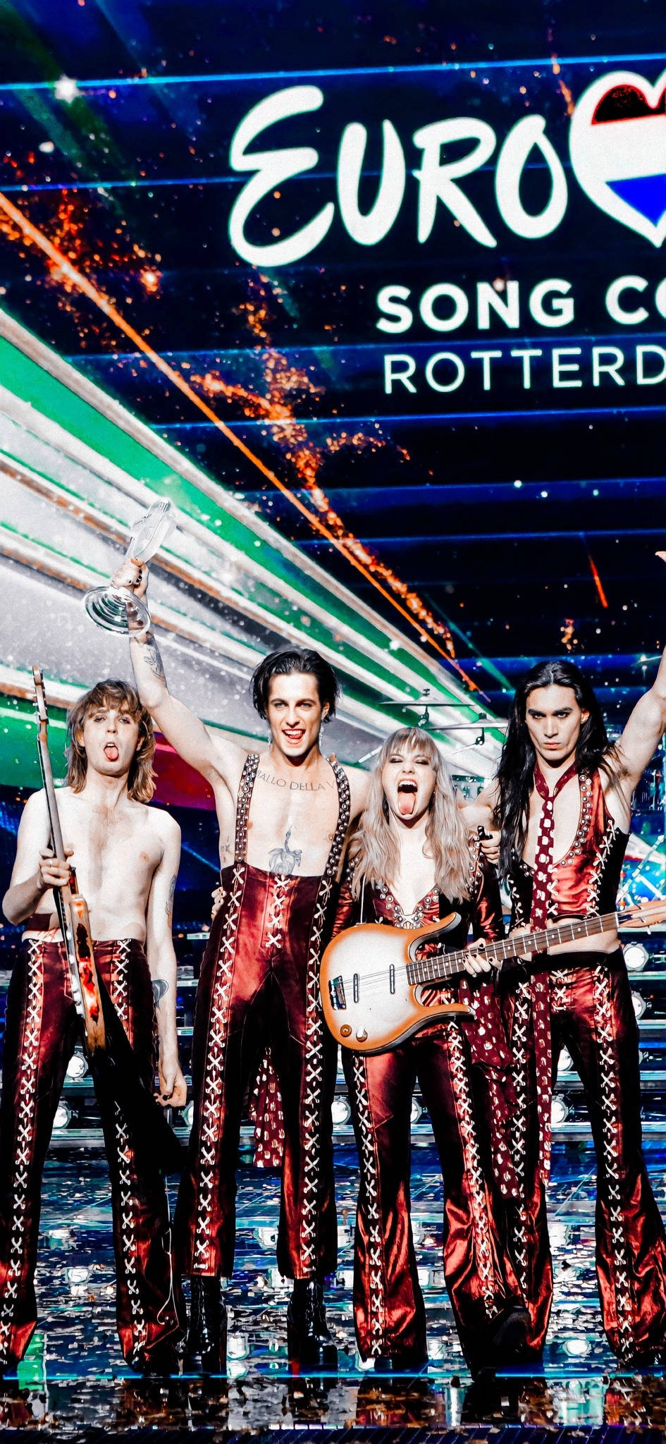 Maneskin 舞台合影 壁紙 — 四位充滿活力的表演者身穿閃亮紅色服裝，在閃閃發光的歐洲歌唱大賽舞台上，手持獎盃和結他慶祝。
