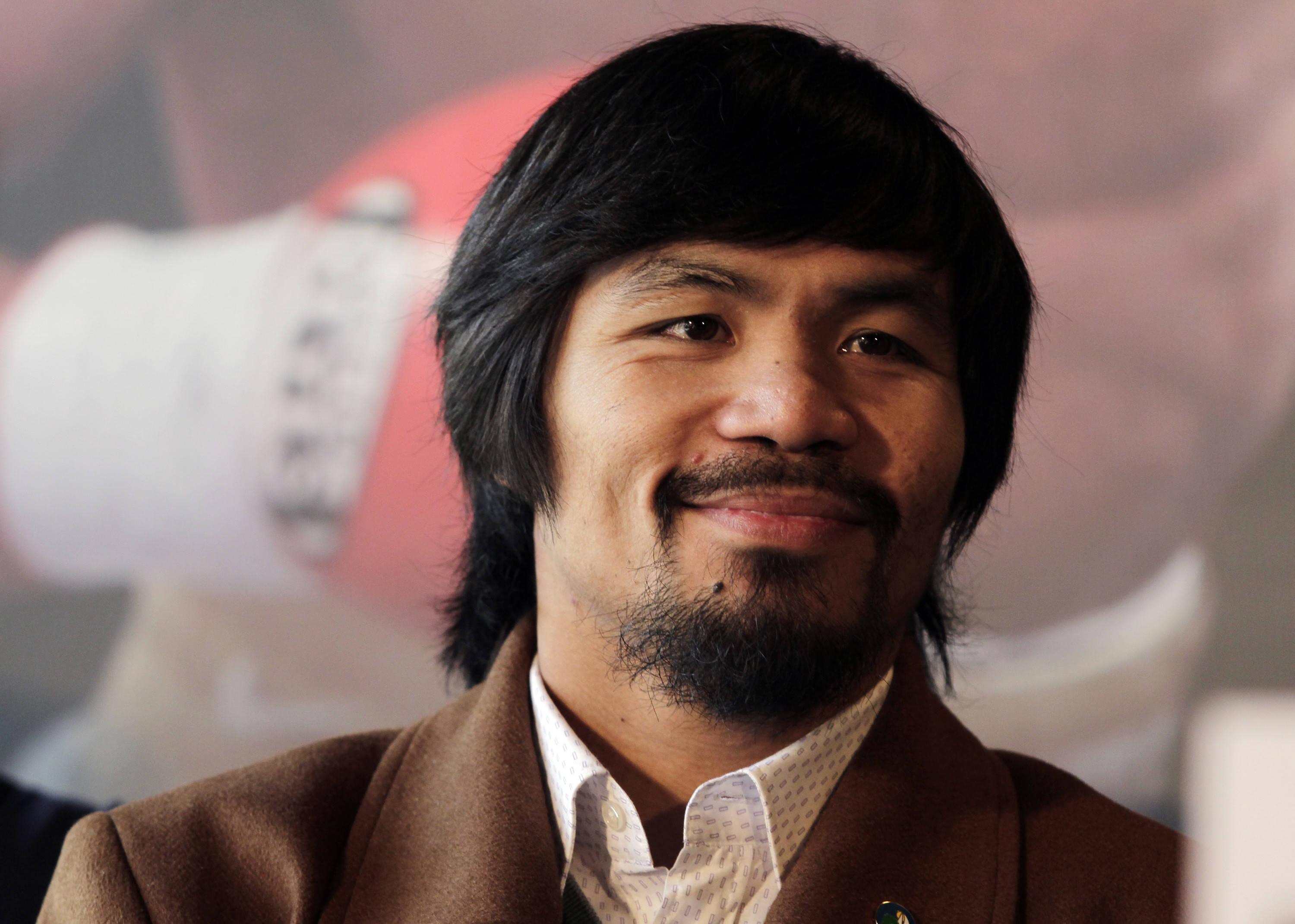 Manny Pacquiao Out Of The Ring 壁紙 — Manny Pacquiao 嘅特寫肖像，佢著住棕色外套，喺模糊嘅拳擊主題背景前溫暖咁微笑。