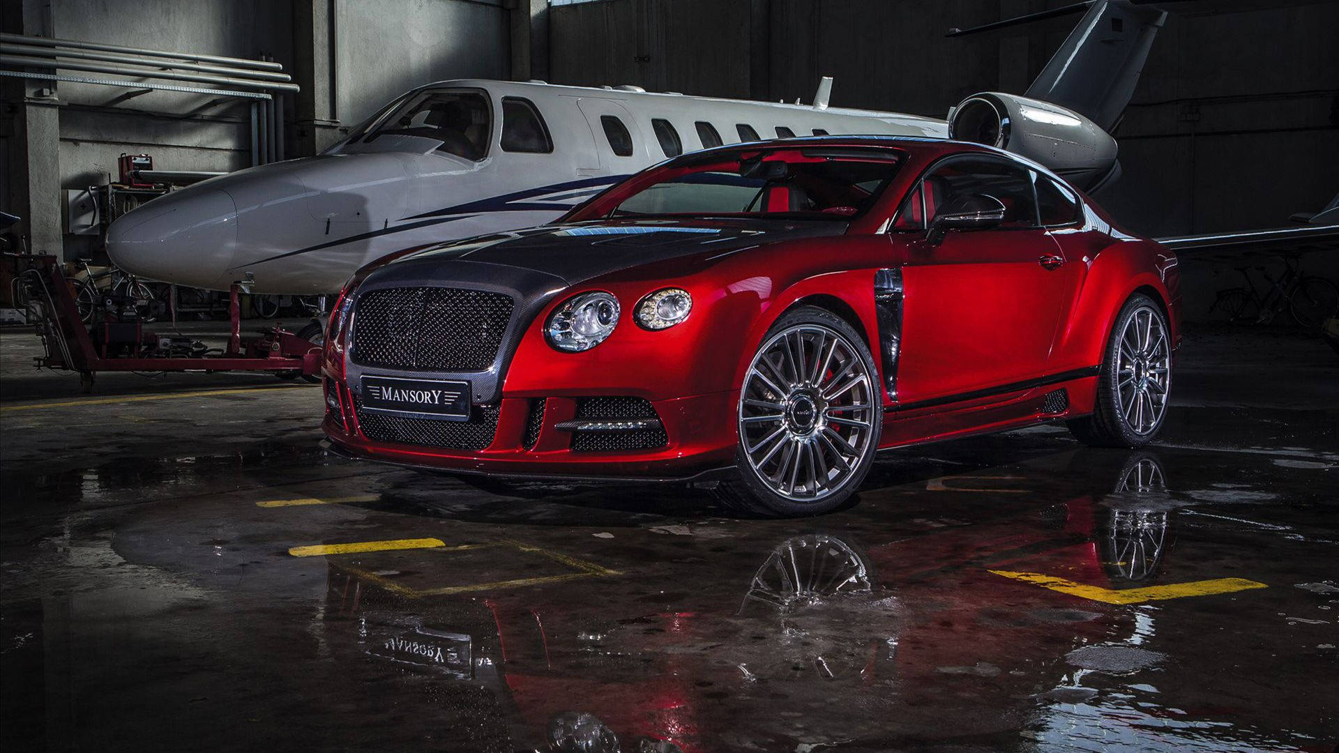 炫目紅色Mansory賓利Continental 壁紙 — 一架亮紅色 Mansory Bentley Continental GT 停泊在潮濕的機庫內，旁邊有一架私人飛機，地板上有倒影。
