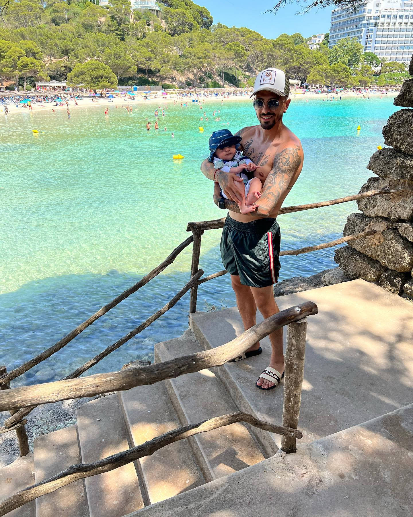 Manuel Lanzini 與嬰兒 壁紙 — 一個微笑的紋身男子在石階上抱著嬰兒，俯瞰著充滿活力的碧綠海灘，人們在游泳，背景是鬱鬱蔥蔥的綠色山丘。