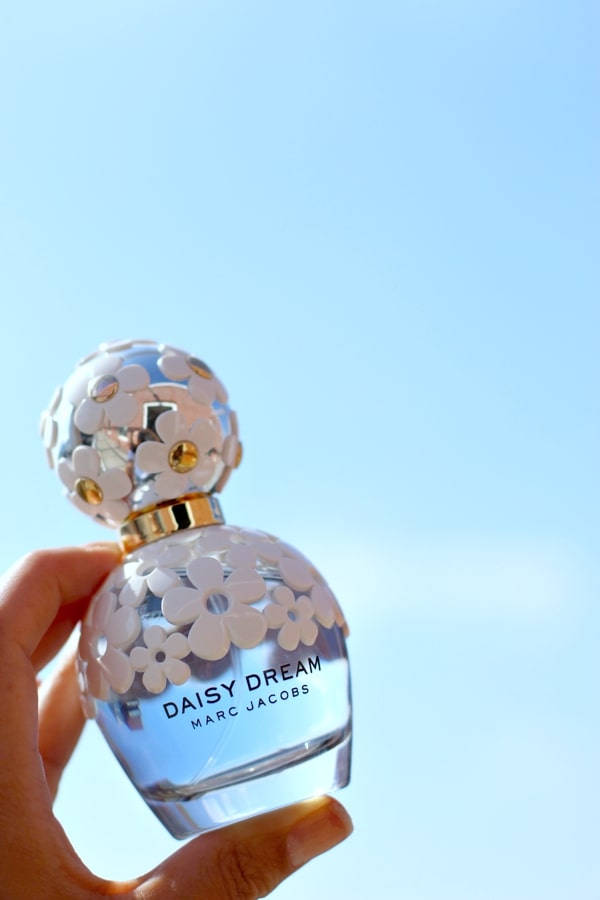 Marc Jacobs Daisy Dream 香水 壁紙 — 一隻手拿著雛菊主題香水瓶，背景是清澈的藍天，陽光在花朵形狀的瓶蓋上閃爍。