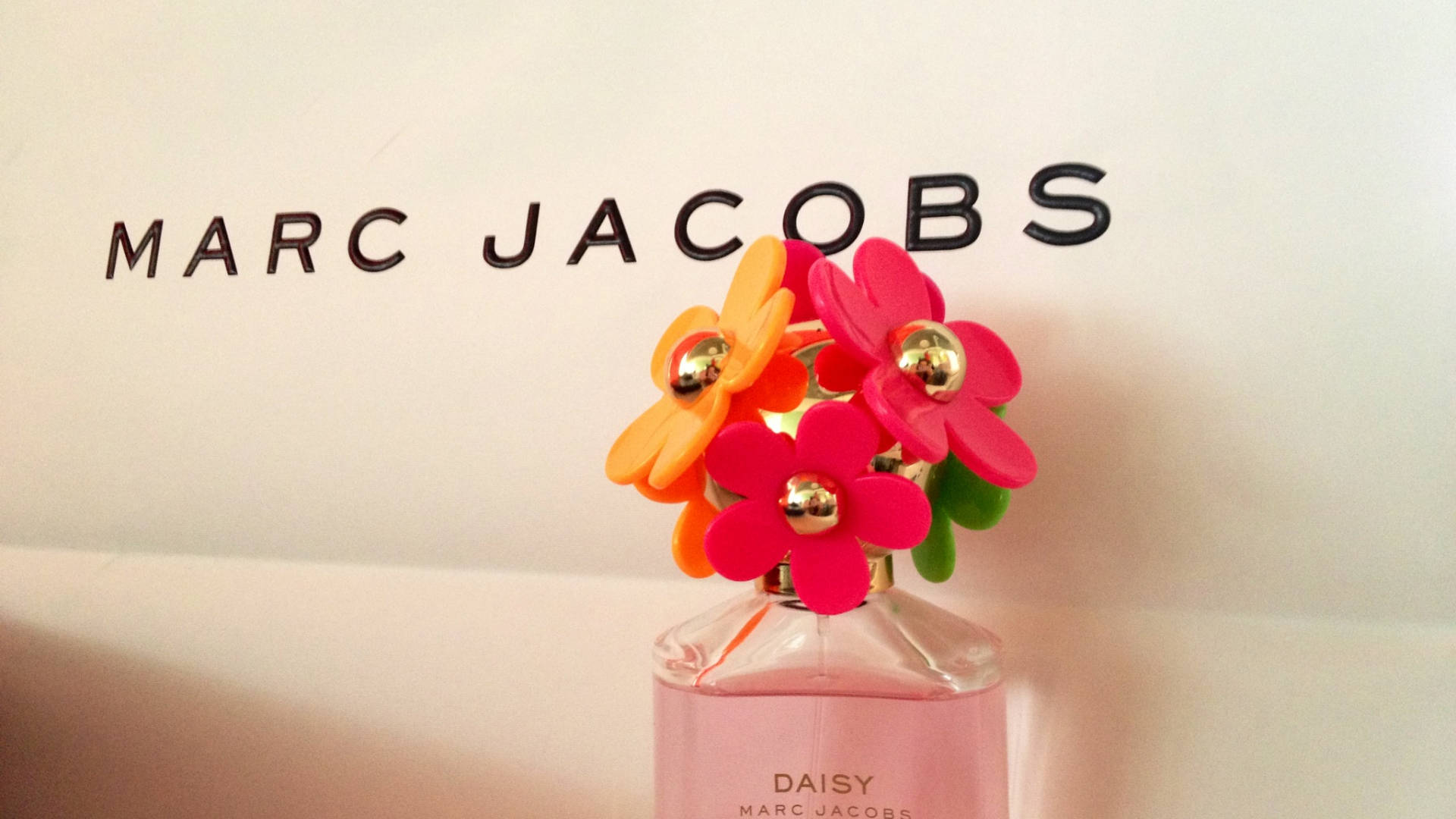 Marc Jacobs 小蒼蘭清新香氛 壁紙 — 一個粉紅色的 Marc Jacobs Daisy 香水瓶，頂部飾有鮮橙色和粉紅色的塑膠花朵，靠著一面印有黑色品牌名稱的白牆。