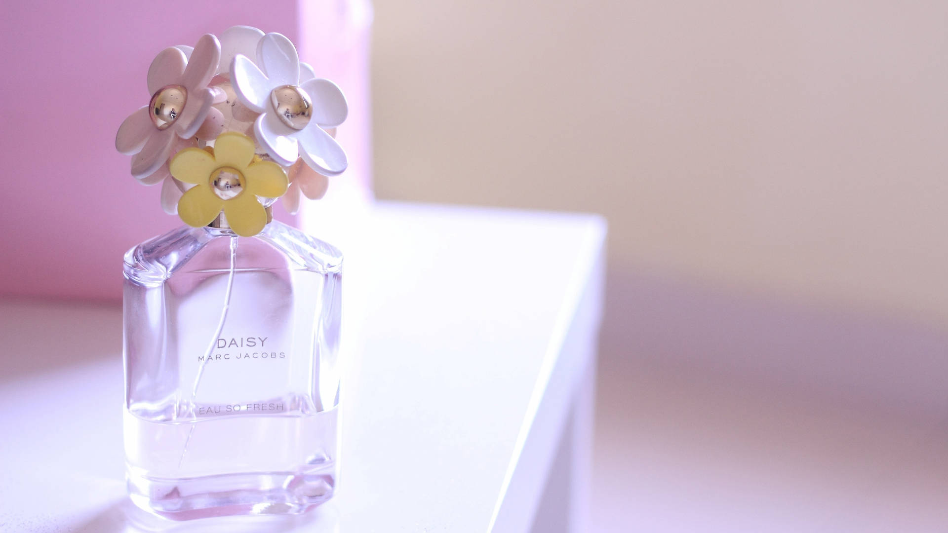 Marc Jacobs 小蒼蘭香氛瓶 壁紙 — 一個透明玻璃香水瓶，標有 Marc Jacobs 的 Daisy，頂部有粉彩花朵裝飾，放置在白色表面上，背景為柔和粉紅色。