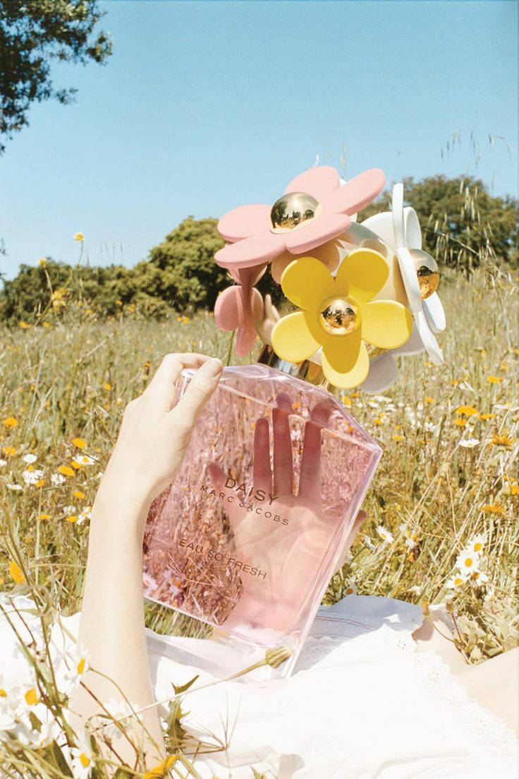 Marc Jacobs 秘密花園香氛版 壁紙 — 一隻手在陽光明媚的野花田中拿著一個帶有花卉裝飾的粉紅色香水瓶。