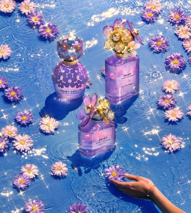 Marc Jacobs 精緻小蒼蘭香氛套裝 壁紙 — 三個紫色香水瓶漂浮在閃爍的藍色水中，周圍環繞著雛菊，一隻手輕輕觸碰花朵。