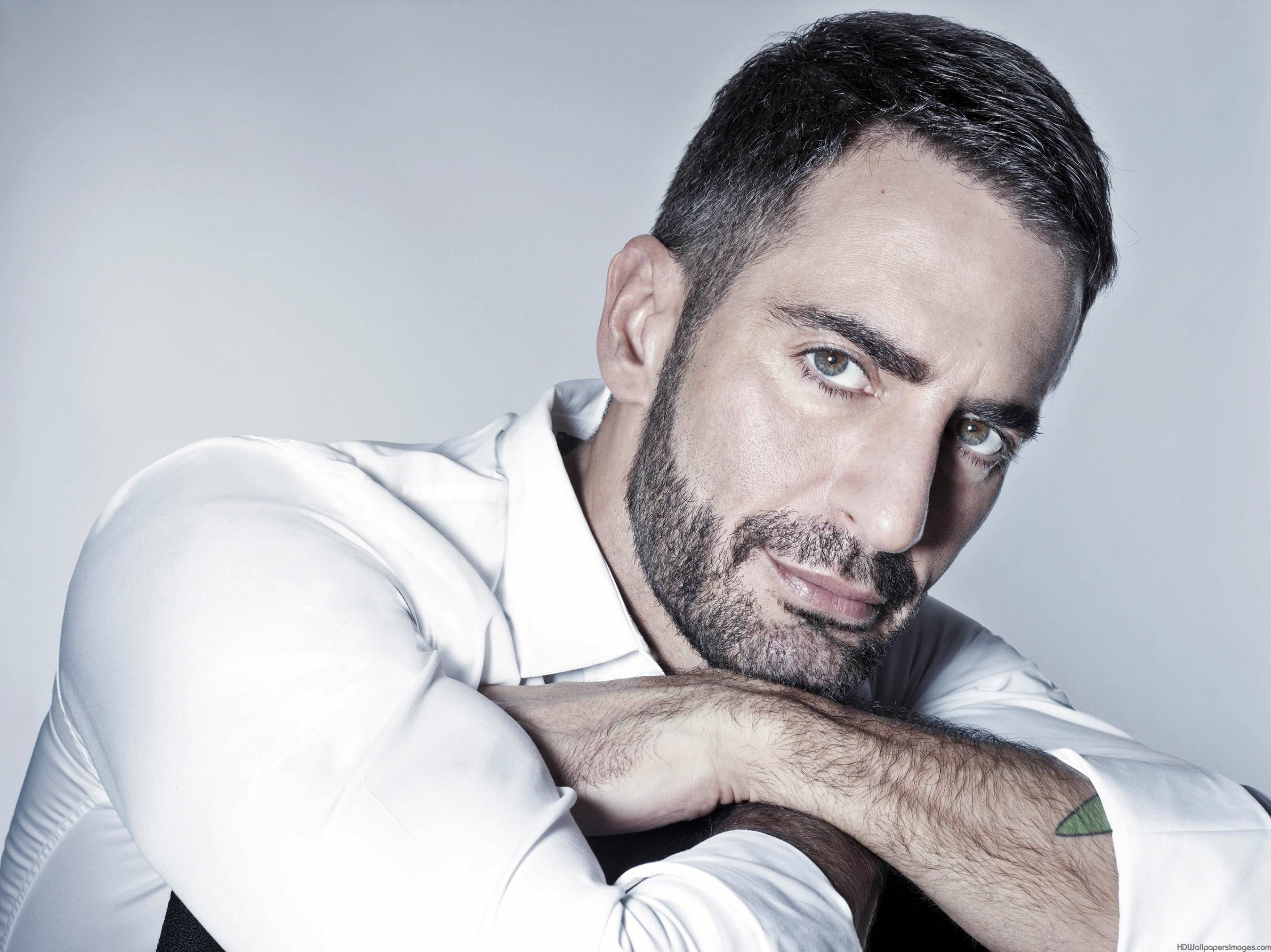 Marc Jacobs創辦人 壁紙 — 一個留鬍鬚、眼神銳利的男人，將下巴擱在手臂上，背景是純灰色，身穿一件挺括的白襯衫。