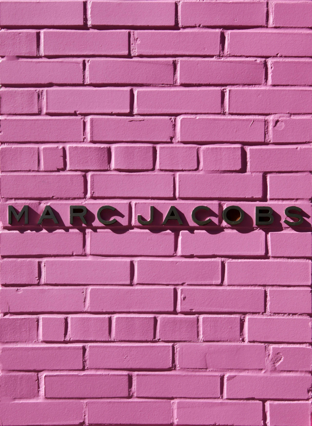 Marc Jacobs粉紅磚牆 壁紙 — 大膽的黑色 'MARC JACOBS' 字母安裝在充滿活力的粉紅色油漆磚牆上。
