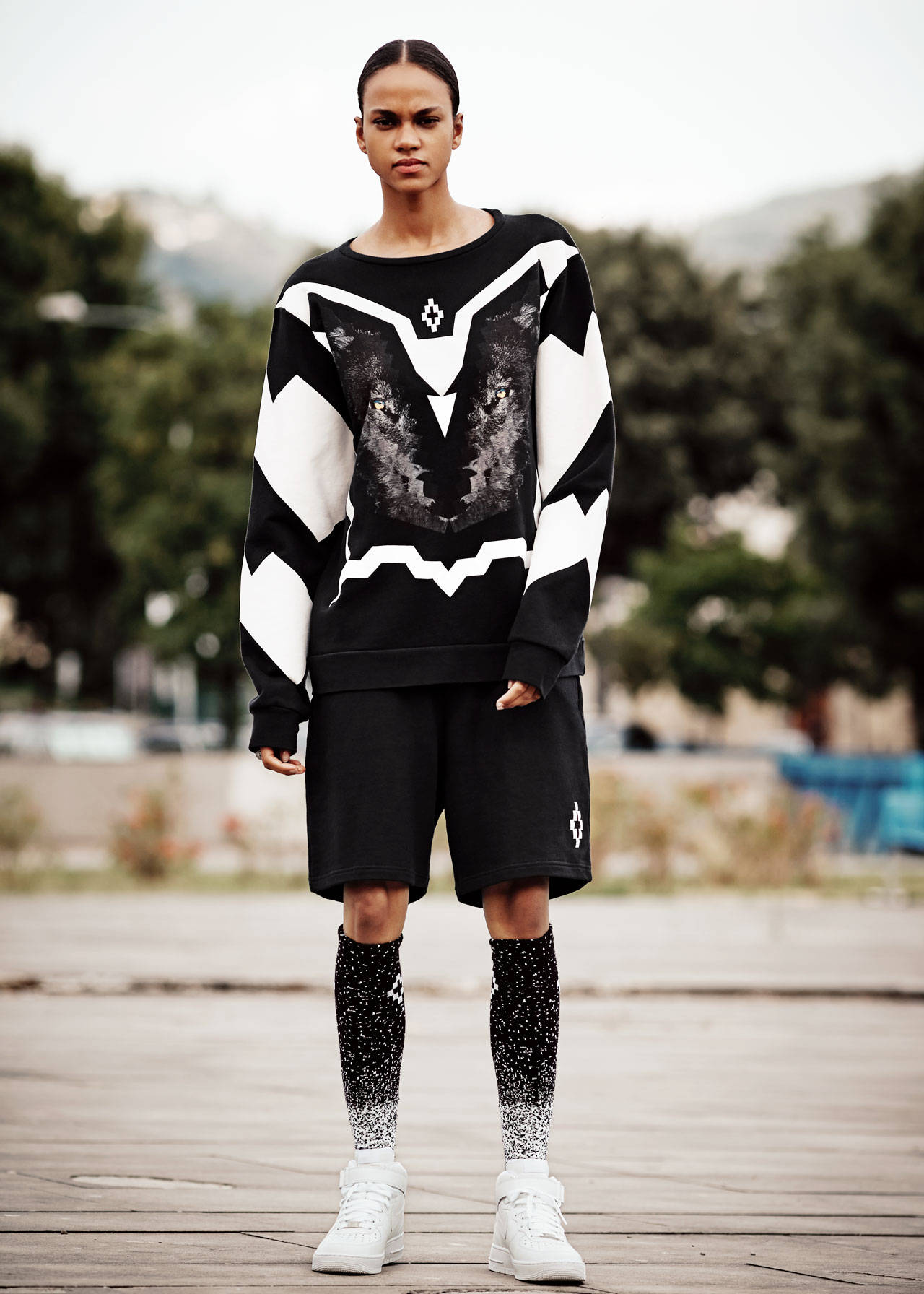 Marcelo Burlon 狼頭賽道圓領衫 壁紙 — 模特兒自信地站在木甲板上，身穿大膽的黑白幾何圖案毛衣，帶有狼圖案，搭配黑色短褲和圖案襪子。