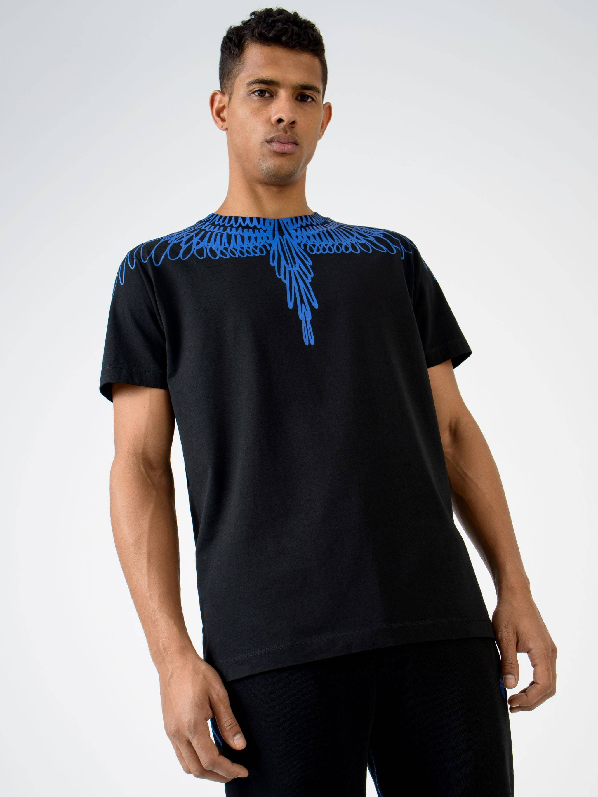 Marcelo Burlon 藍色翅膀印花T恤 壁紙 — 一名男子身穿黑色T恤，肩上有大膽的藍色翅膀圖案，背景為純白色。