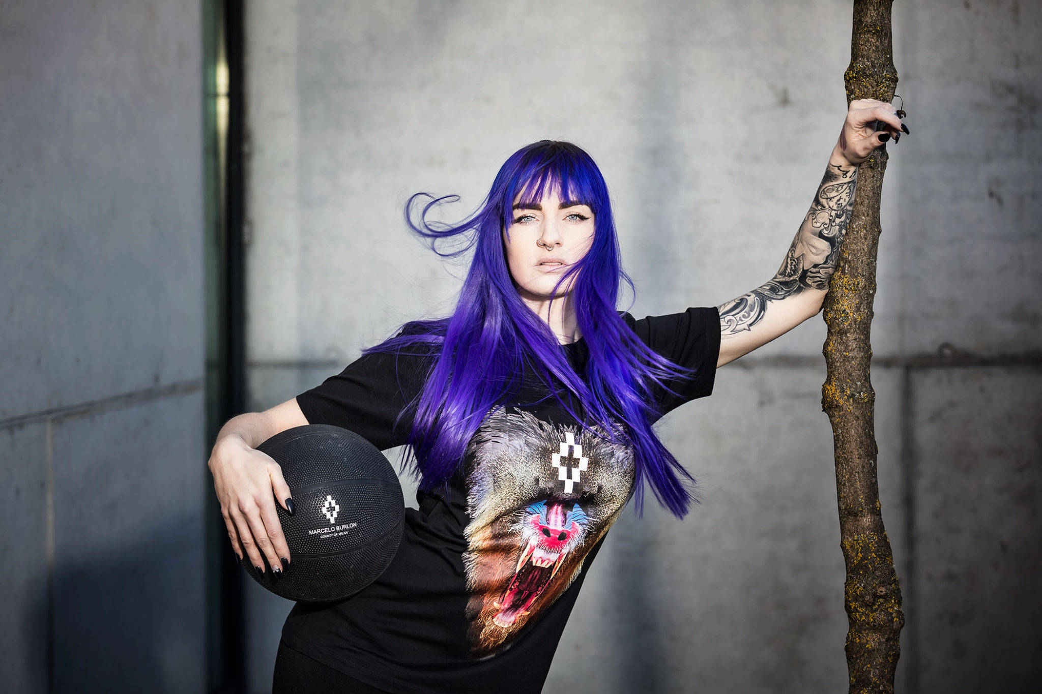 Marcelo Burlon 狂怒動物頭T恤 壁紙 — 一位擁有鮮豔紫色頭髮和紋身的女性靠著樹，將一個黑色球抵在混凝土牆上。