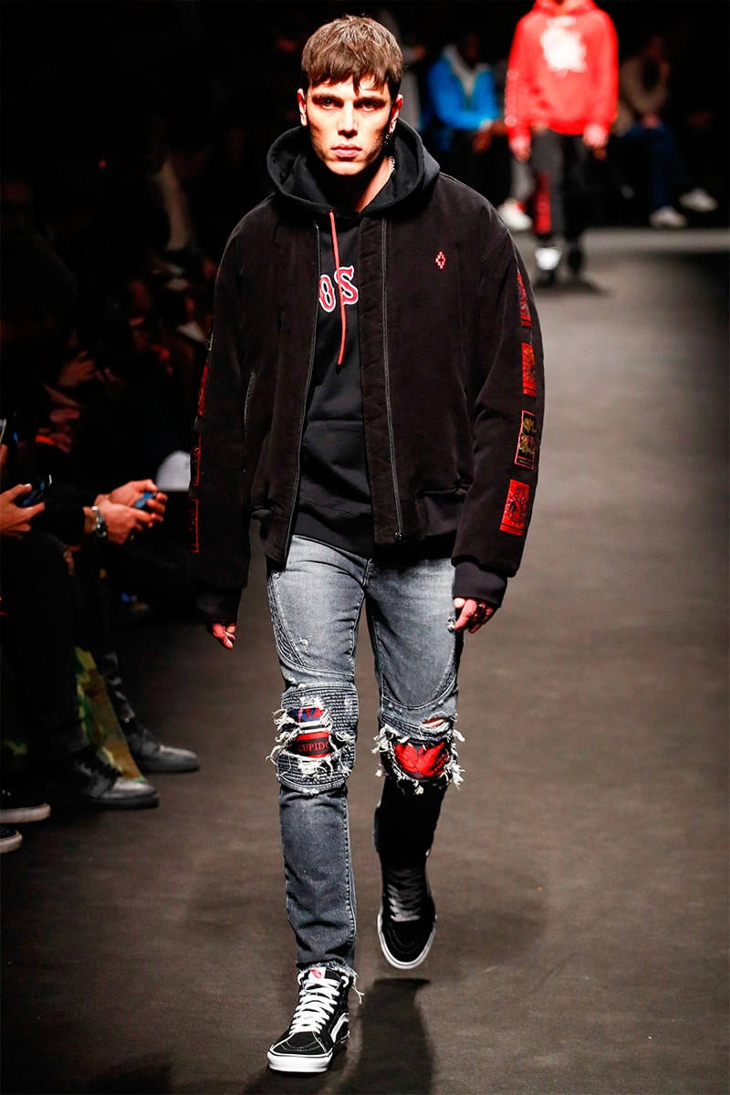 Marcelo Burlon 獨特藝術設計 壁紙 — 一名男模特兒穿著黑色連帽外套、破洞牛仔褲和黑色運動鞋，在明亮的舞台燈光下走秀。