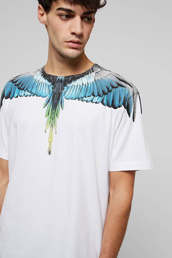 Marcelo Burlon 白色T恤配藍色翅膀 壁紙 — 一名男子穿著白色T恤，上面有生動的藍色和綠色翅膀圖案，橫跨他的肩膀。