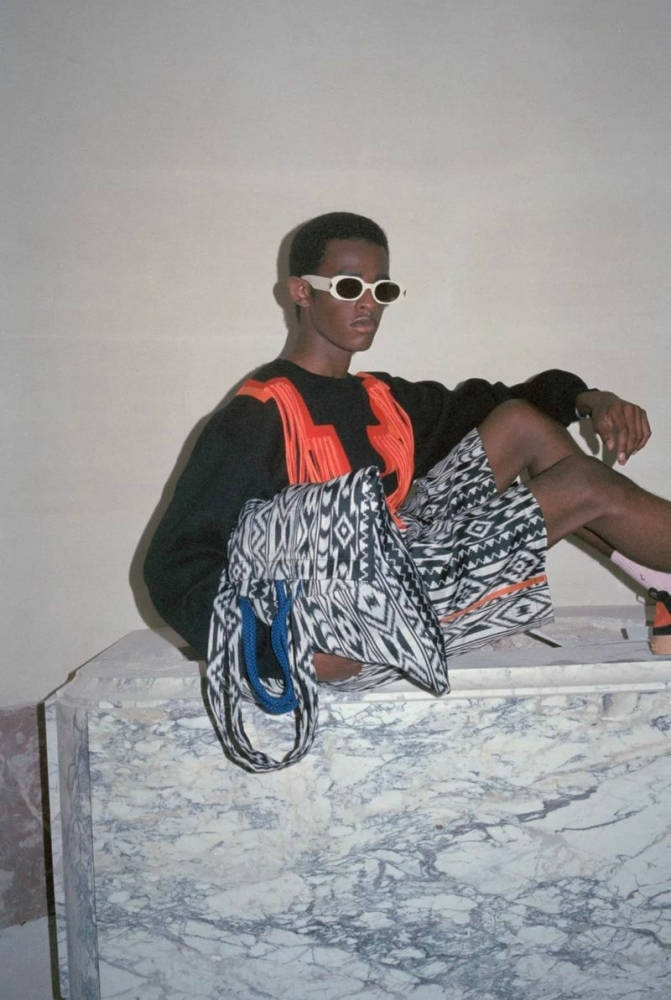 Marcelo Burlon 素色衛衣與圖案短褲 壁紙 — 一位時尚人士戴著白色太陽眼鏡和鮮明圖案服裝，自信地坐在大理石基座上，背景是素牆。