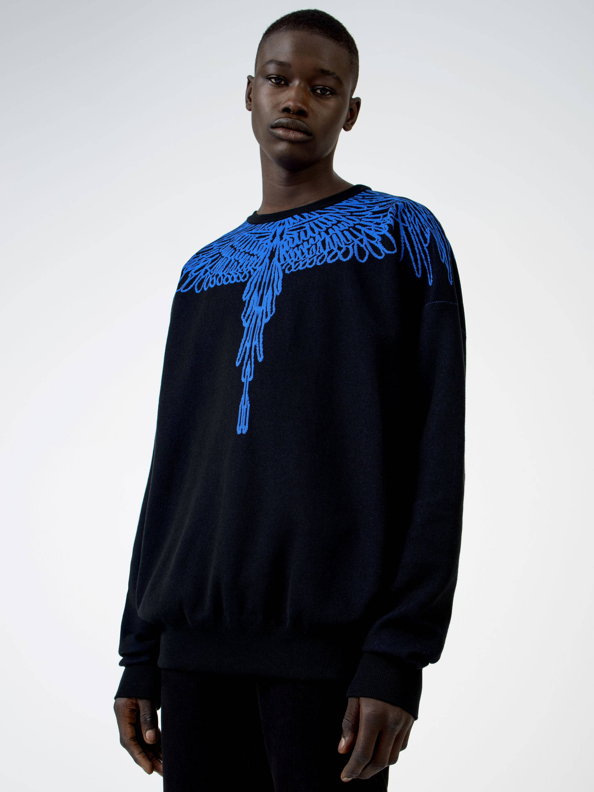 Marcelo Burlon 藍色翅膀印花連帽衫 壁紙 — 一名身穿黑色毛衣、肩上帶有醒目藍色刺繡翅膀圖案的男人，站在純白色背景前。