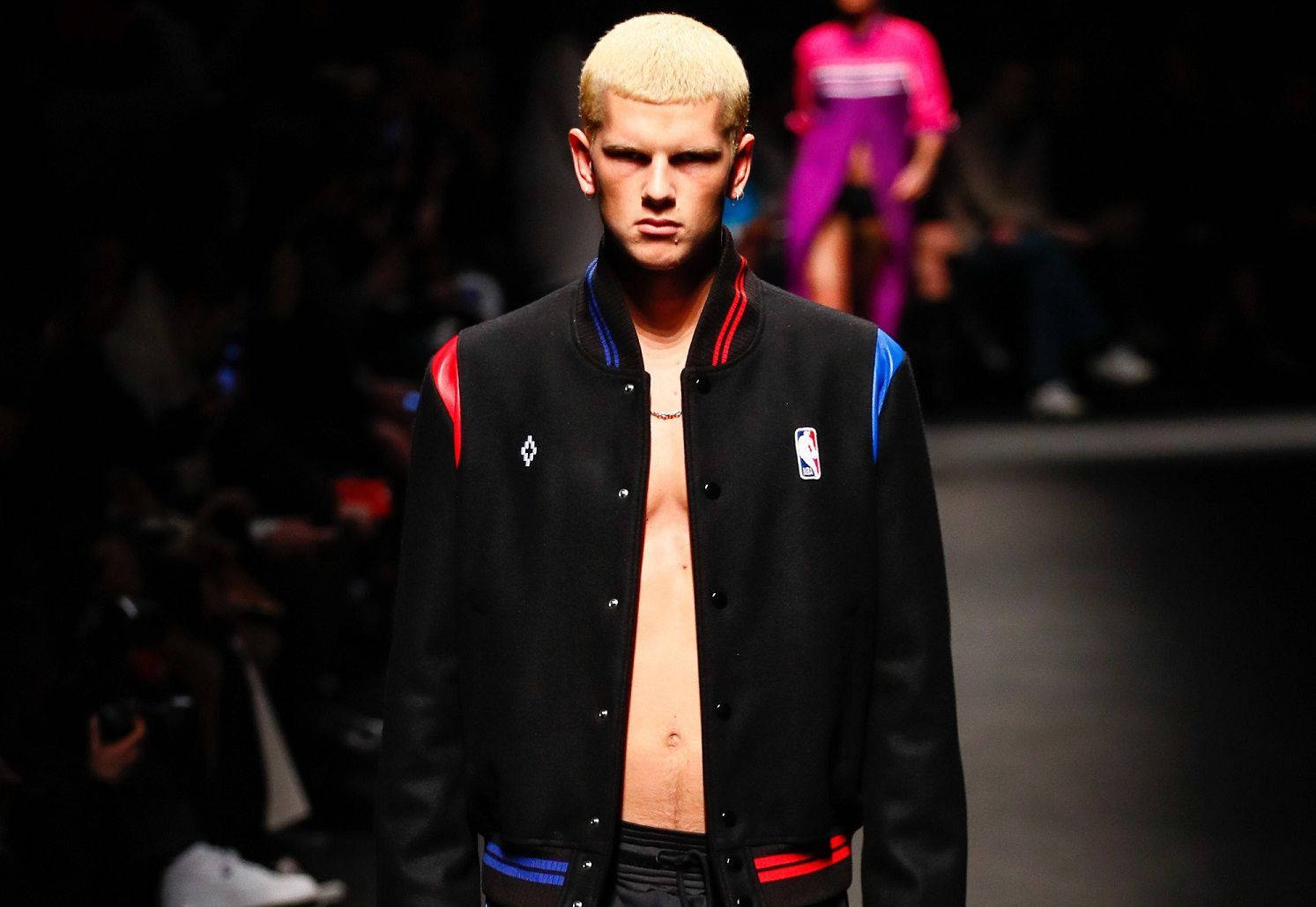 Marcelo Burlon 黑色飛行員夾克 - 經典與都市風格的化身 壁紙 — 一位擁有鉑金色頭髮的模特兒自信地走在黑暗的T台上，身穿一件帶有紅藍色點綴和NBA標誌的黑色開襟Varsity外套。