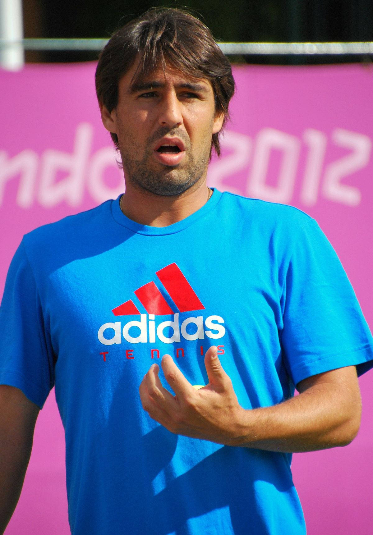marcos_baghdatis_穿_adidas_藍色上衣 壁紙 — 一名身穿藍色adidas網球衫的男子在粉紅色2012背景下說話時做手勢。