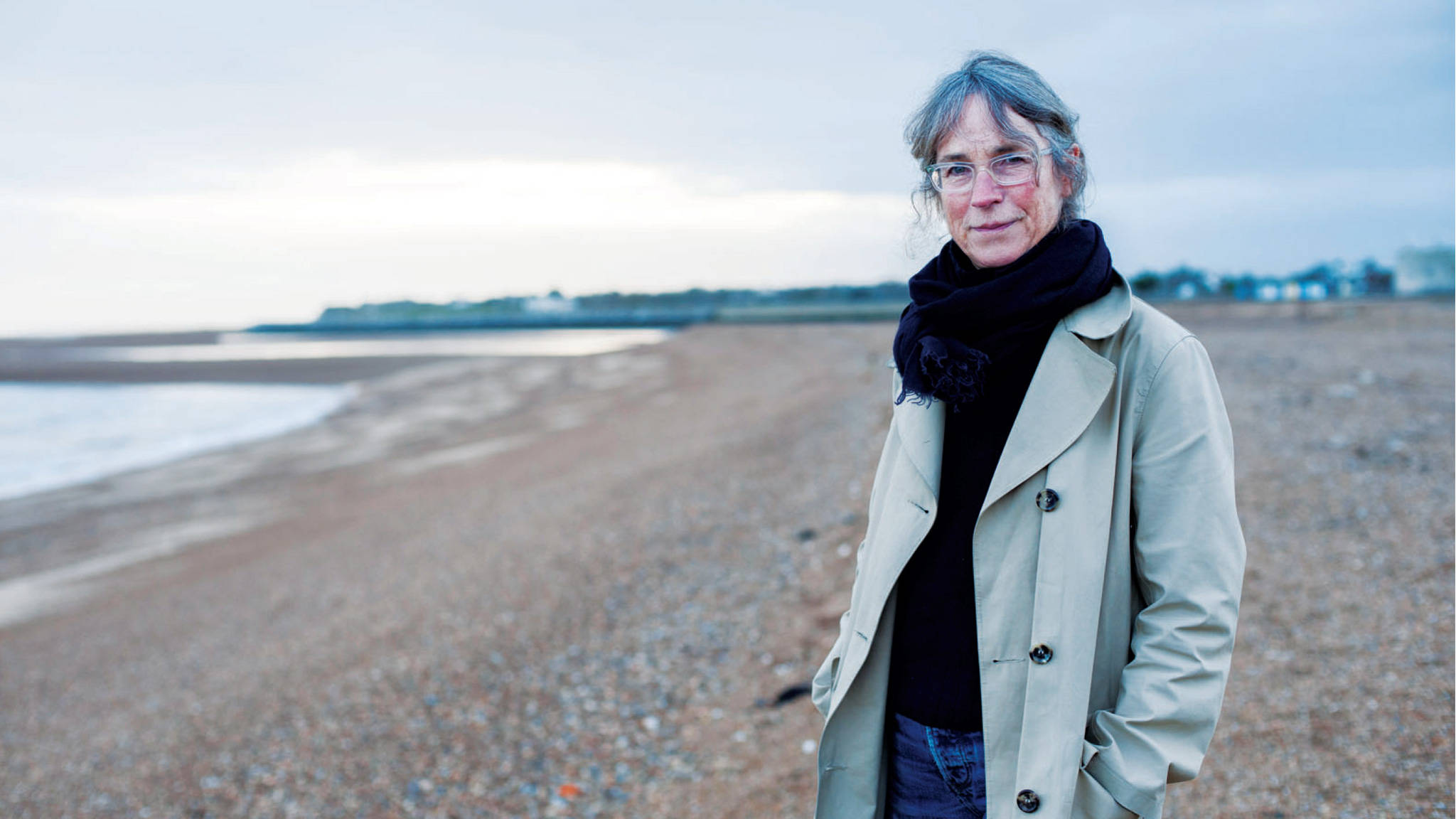 Margaret Howell 在寧靜海灘悠閒漫步 壁紙 — 一位戴眼鏡和圍巾的女性站在卵石海灘上，波浪在陰天輕輕拍打著海岸。