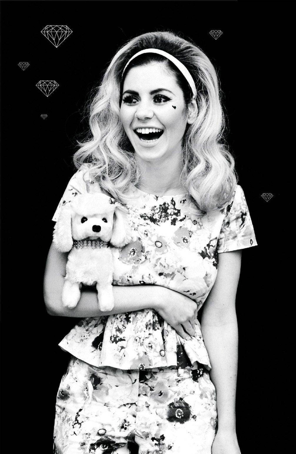 Marina and the Diamonds 拿玩偶 壁紙 — 一位快樂的金色捲髮女子戴著頭帶，抱著一隻毛絨狗，在黑色背景上帶有鑽石輪廓的畫面中大笑。