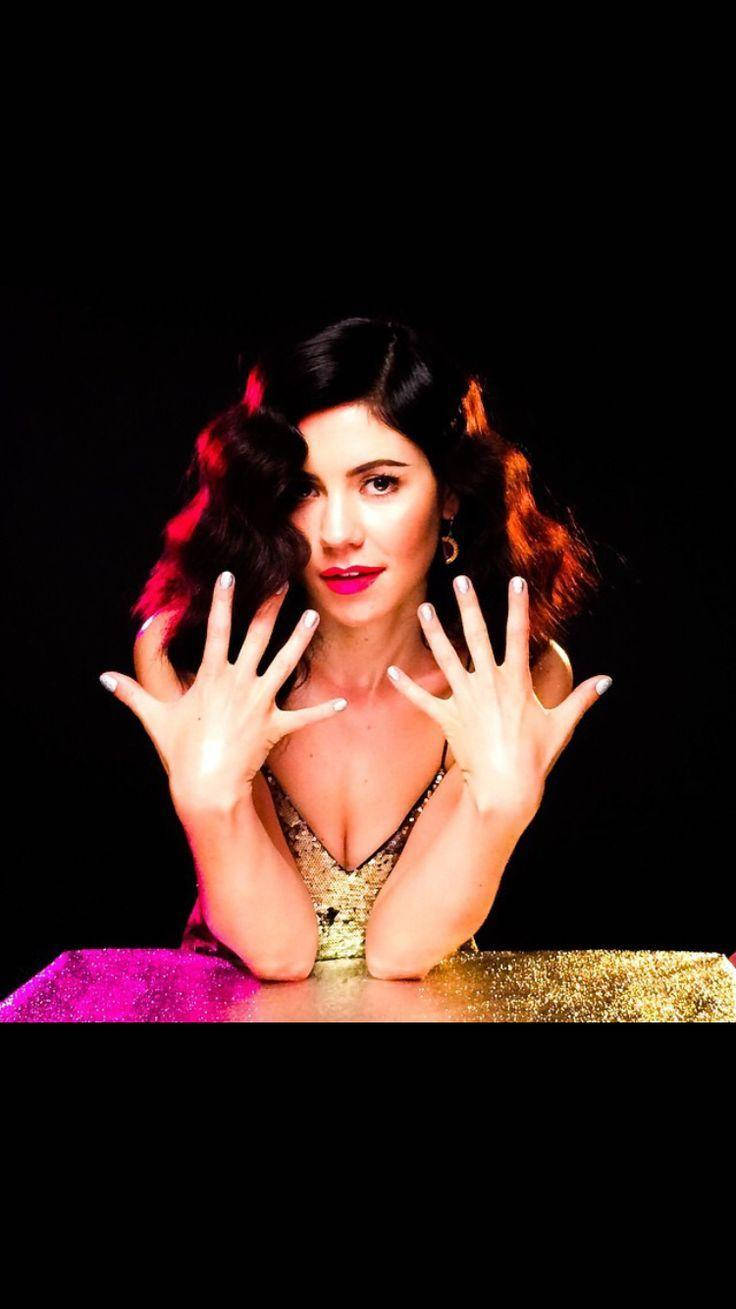 Marina and the Diamonds 美麗造型 壁紙 — 一位擁有深色波浪頭髮和大膽紅唇的女性，在黑色背景上，雙手放在閃閃發光的粉紅色和金色表面上擺姿勢。