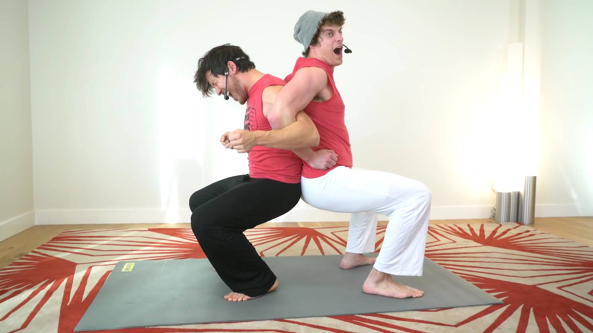 Markiplier 朋友瑜珈 壁紙 — 兩名身穿紅色上衣的男子在明亮的房間內，於紅色圖案地毯上的灰色墊子上做出動態瑜伽姿勢。