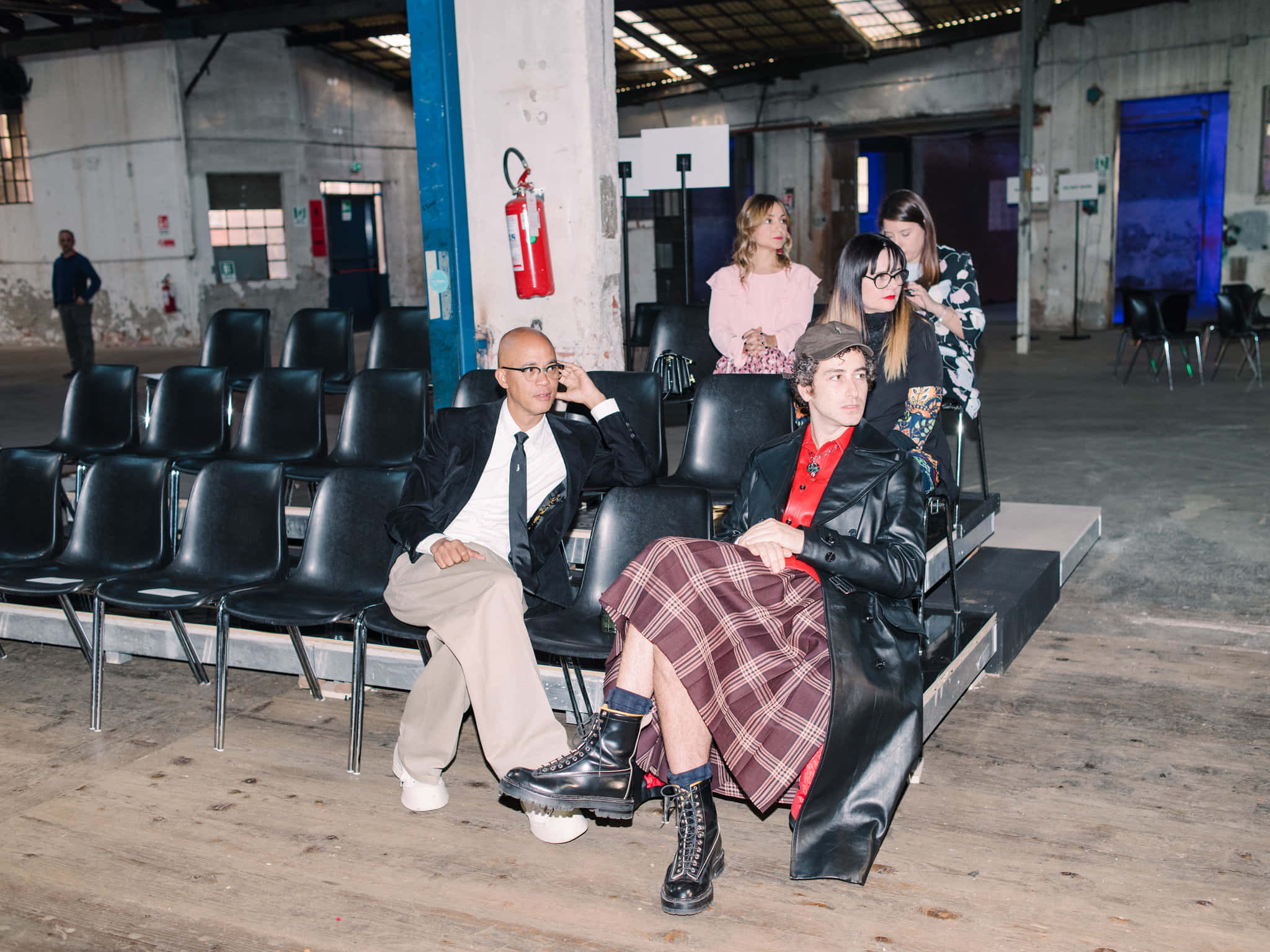 Marni 2019秋冬時裝秀幕後花絮 壁紙 — 四位穿著時尚的人士在一個粗糙的工業場所中擺姿勢，現場有黑色椅子的排列和裸露的混凝土牆壁。
