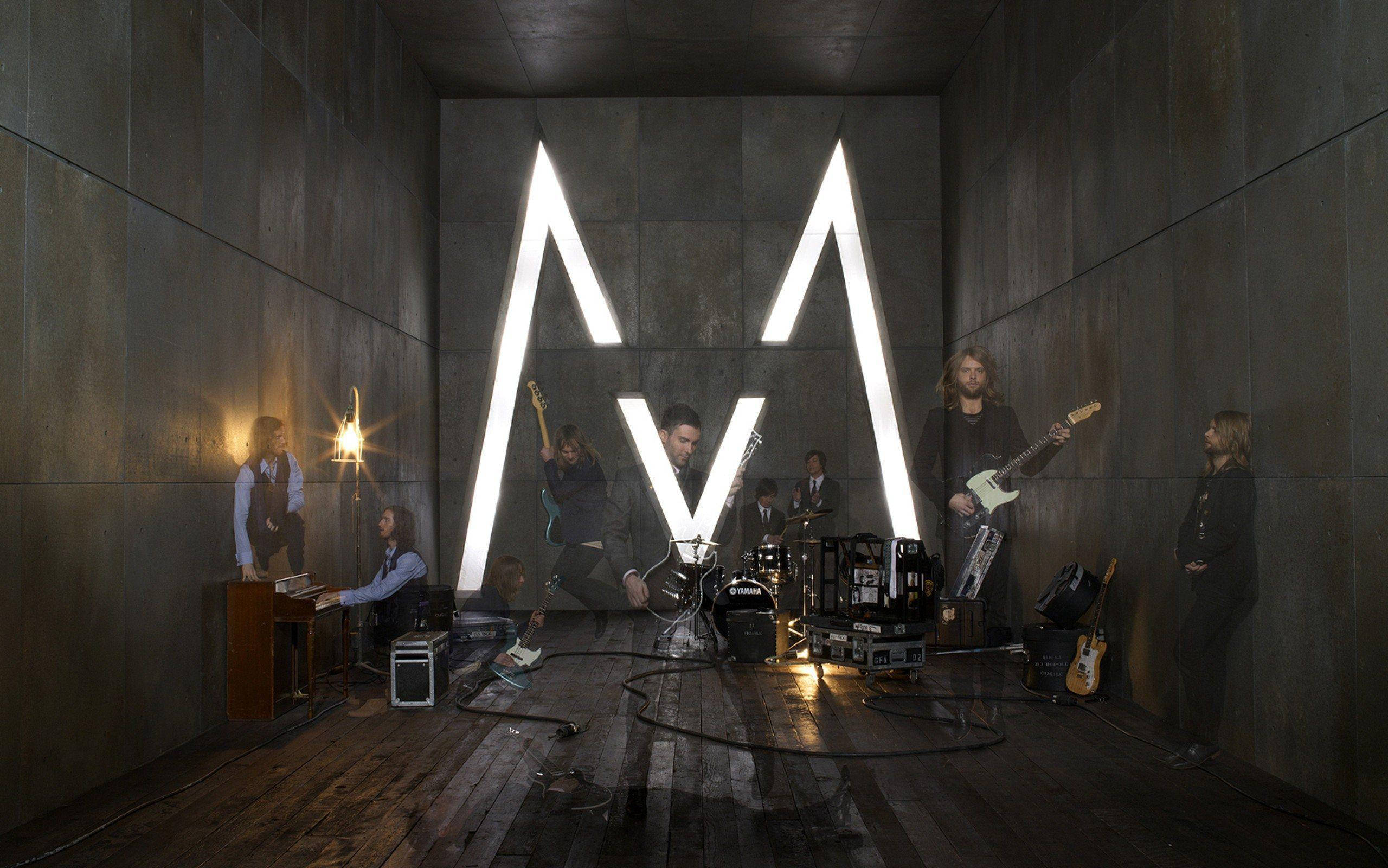 Maroon 5 M 錄音室 壁紙 — 一個樂隊在昏暗的混凝土房間中表演，被後牆上發光的白色「M」標誌照亮。