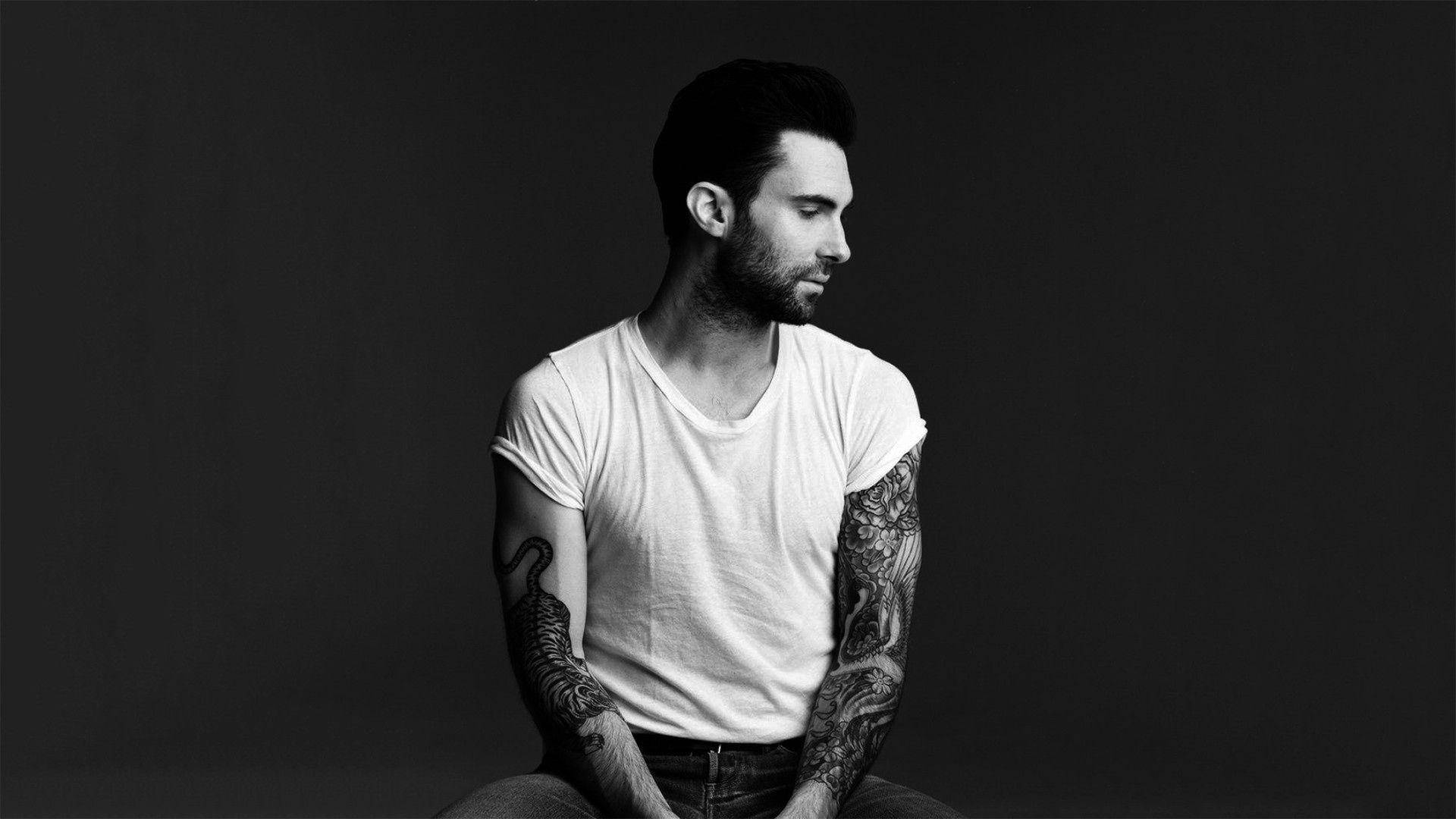 Maroon 5 亞當·列文 黑底白襯衫 壁紙 — 一個有紋身的男人側面坐在深色背景前，穿著白色T恤。