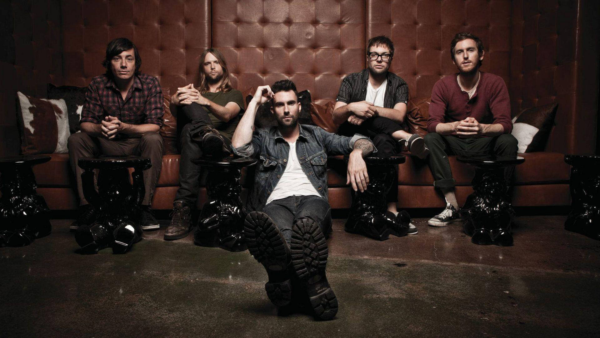 Maroon 5 坐在棕色皮革沙發上 壁紙 — 五位男士穿著休閒服裝，在光線昏暗的休息室裡，自信地坐在簇絨皮革沙發上，地板深色，旁邊有華麗的邊桌。