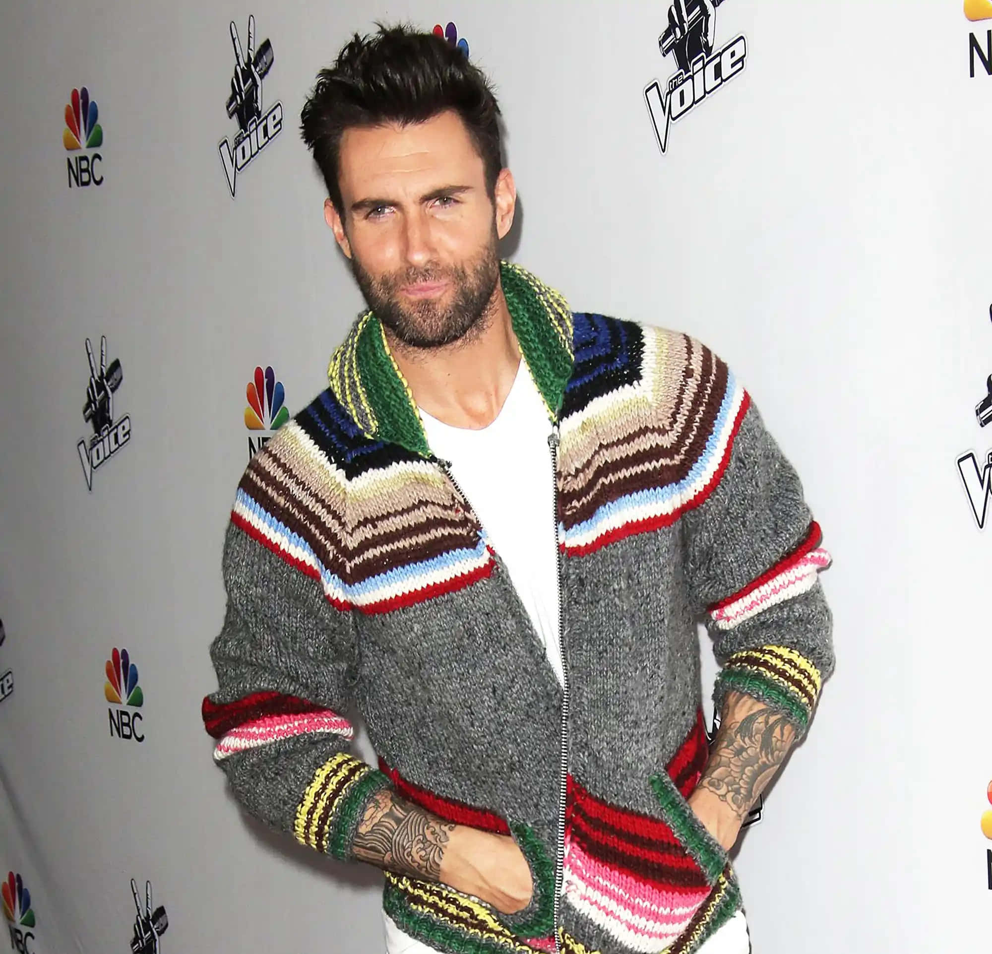 Maroon 5主唱亞當·萊文 壁紙 — Adam Levine 在The Voice活動中自信地擺姿勢，身穿色彩繽紛的條紋針織外套，身後有NBC標誌。